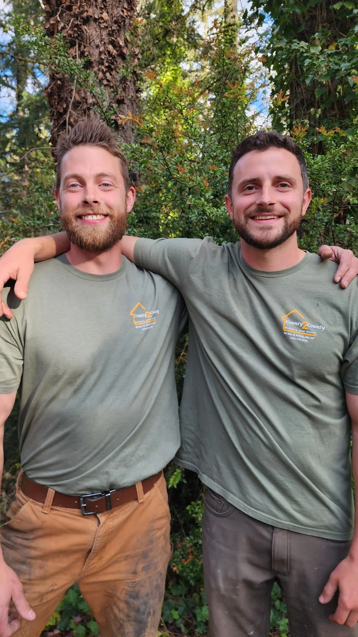 Two men standing outdoors with their arms around each other, smiling. They are wearing matching green T-shirts with a logo that says "County 2 County" and brown and gray pants.. The background features trees and green foliage.