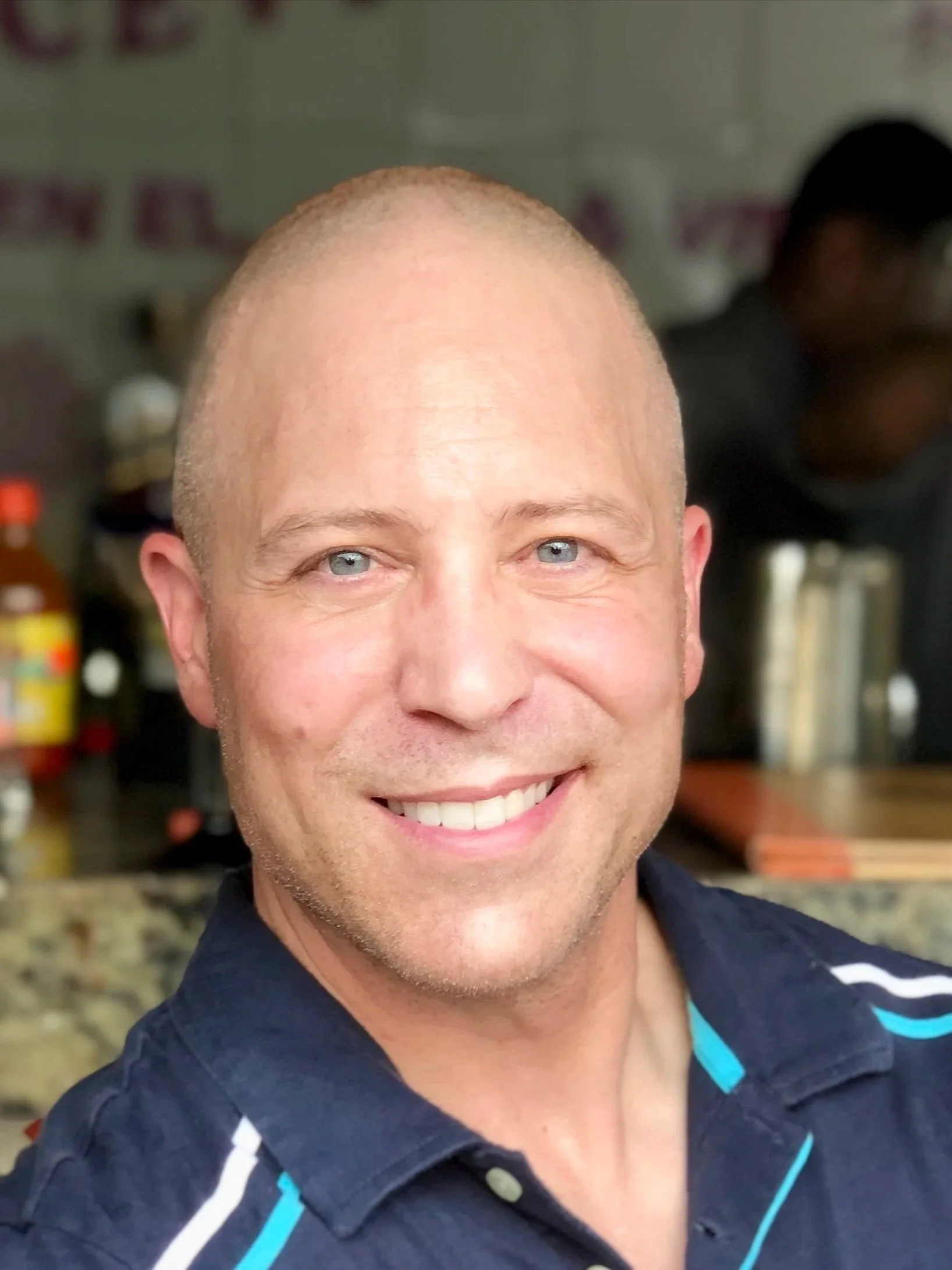 Smiling man with short hair and blue eyes wearing a navy blue collared shirt with light blue and white stripes.