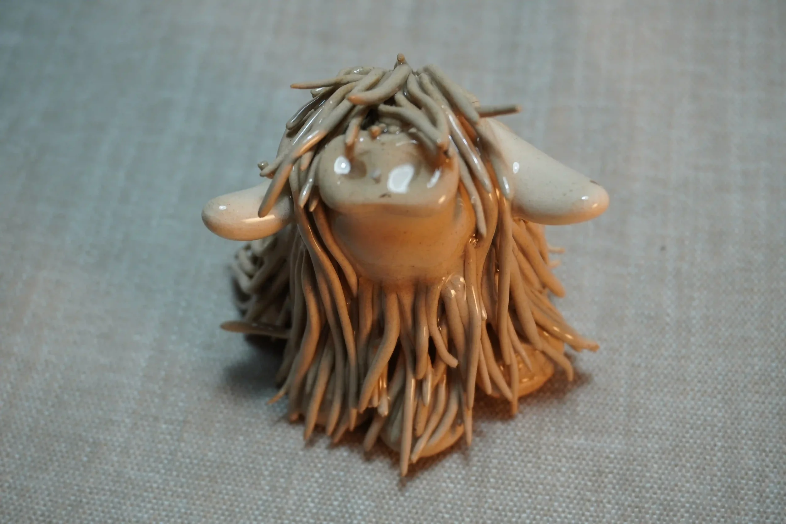 Front view of a handmade ceramic sculpture of a Highland Cow ornament with light tan-coloured hair and beige horns.