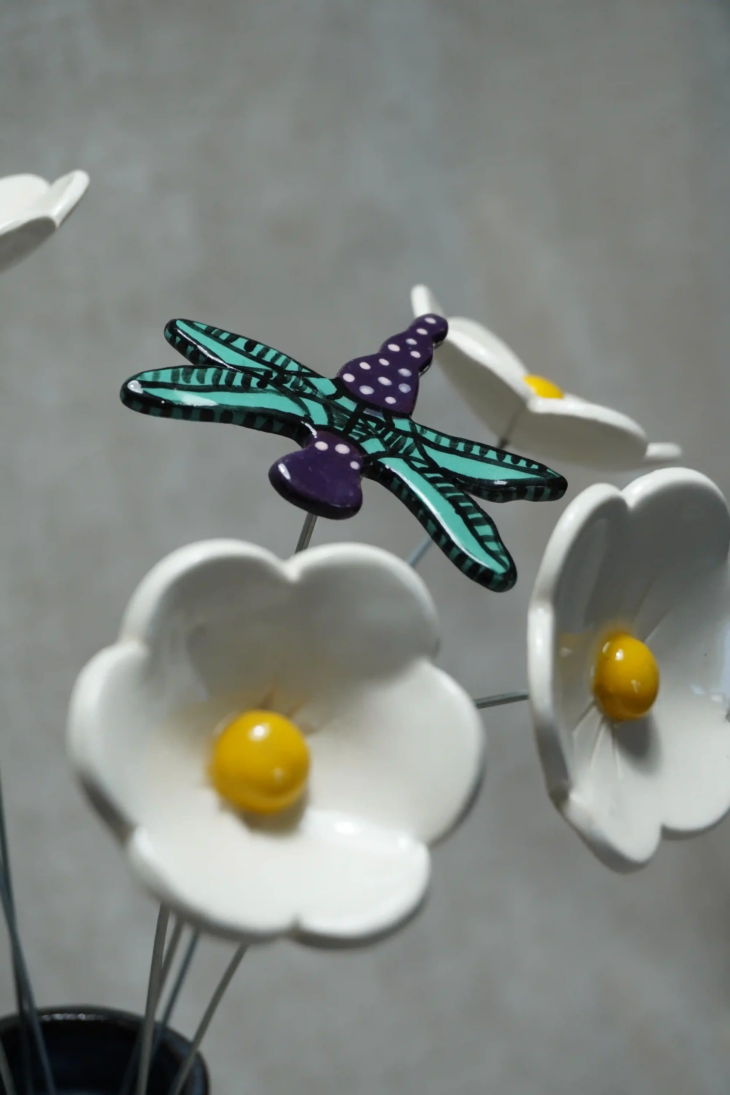 A ceramic dragonfly with purple body with white speckles and teal wings, on a metal stem displayed among white ceramic daisies with yellow centres.