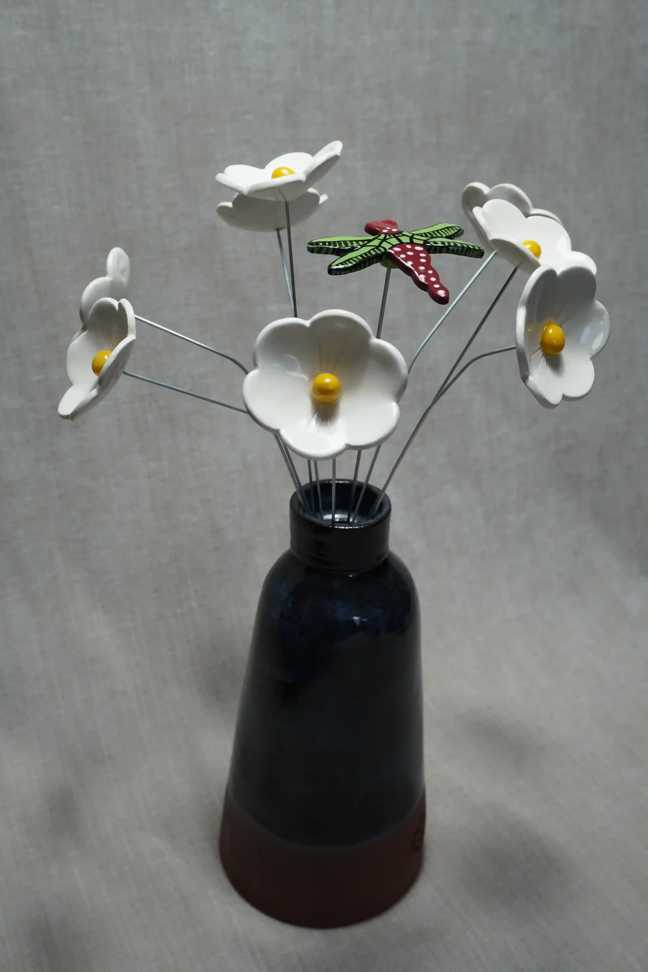 	
A dark blue and terracotta ceramic vase holding ceramic white Daisy flowers with yellow centers, and a colorful ceramic dragonfly with green and black wings and a red body with white speckles, on thin metal stems.