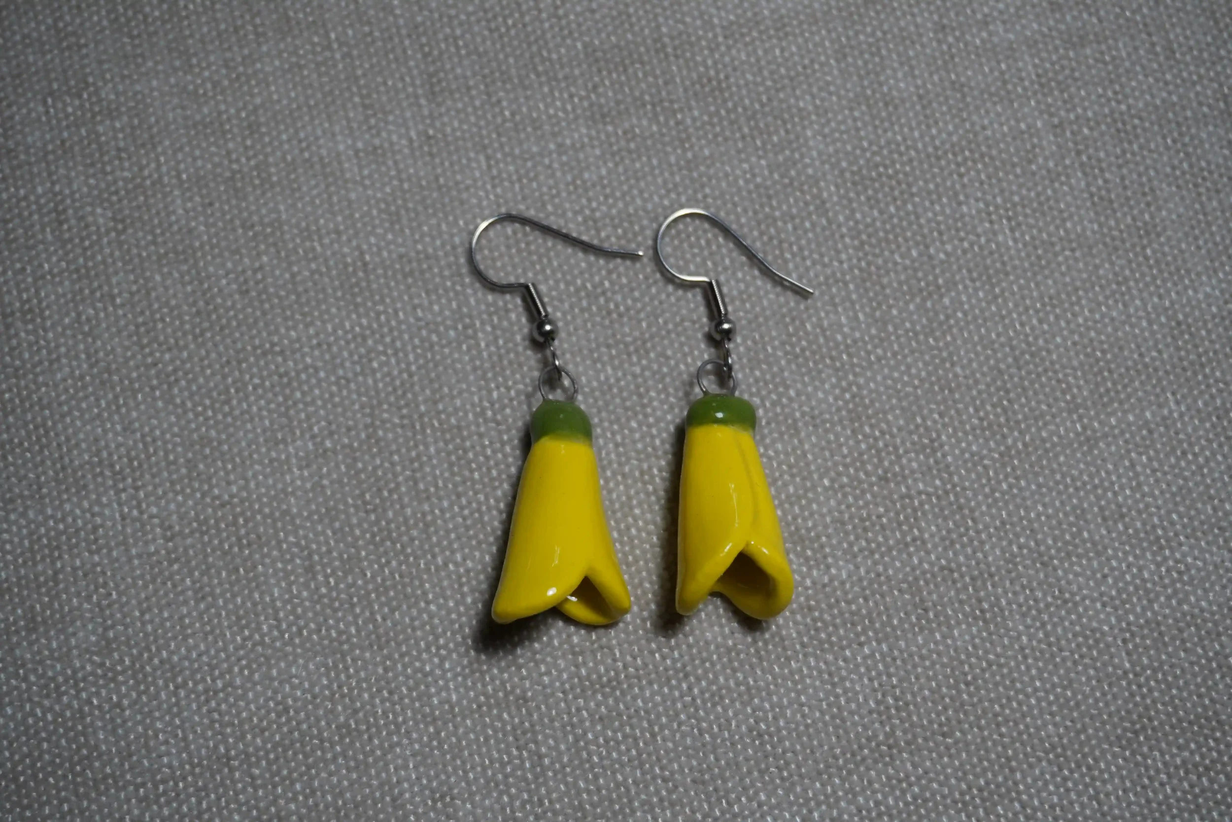 Pair of ceramic earrings with yellow New Zealand Kowhai flower pendants and silver hooks on a textured gray background.