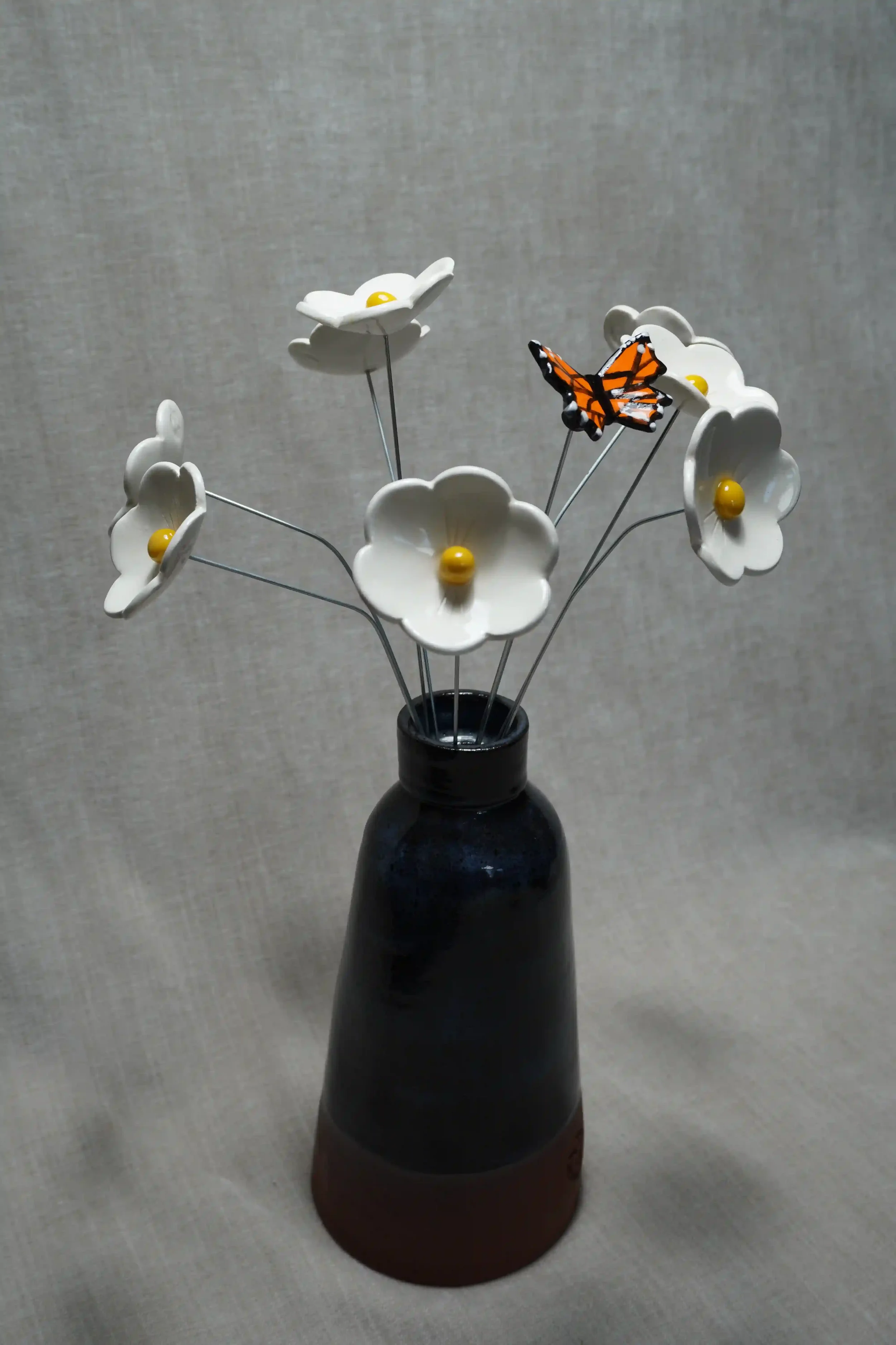 A dark blue-glazed handmade ceramic vase holding white ceramic Daisy flowers with yellow centers and a decorative ceramic Monarch butterfly, set against a neutral background.