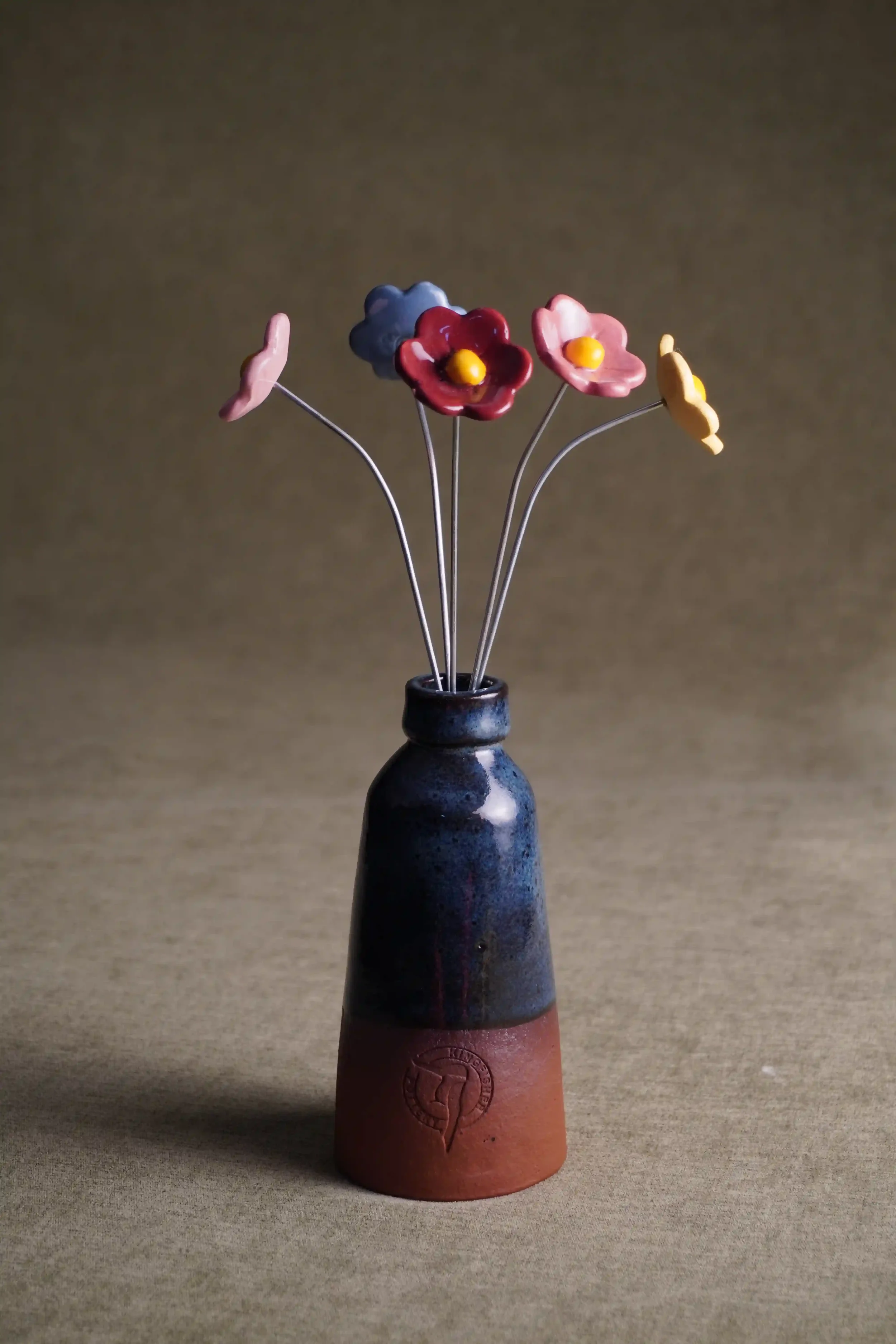 A ceramic vase with a bottle shape, dark blue glaze and terracotta base and prominent mark, holding five ceramic flowers in pink, dark pink, yellow and blue. Shown against a neutral backdrop.