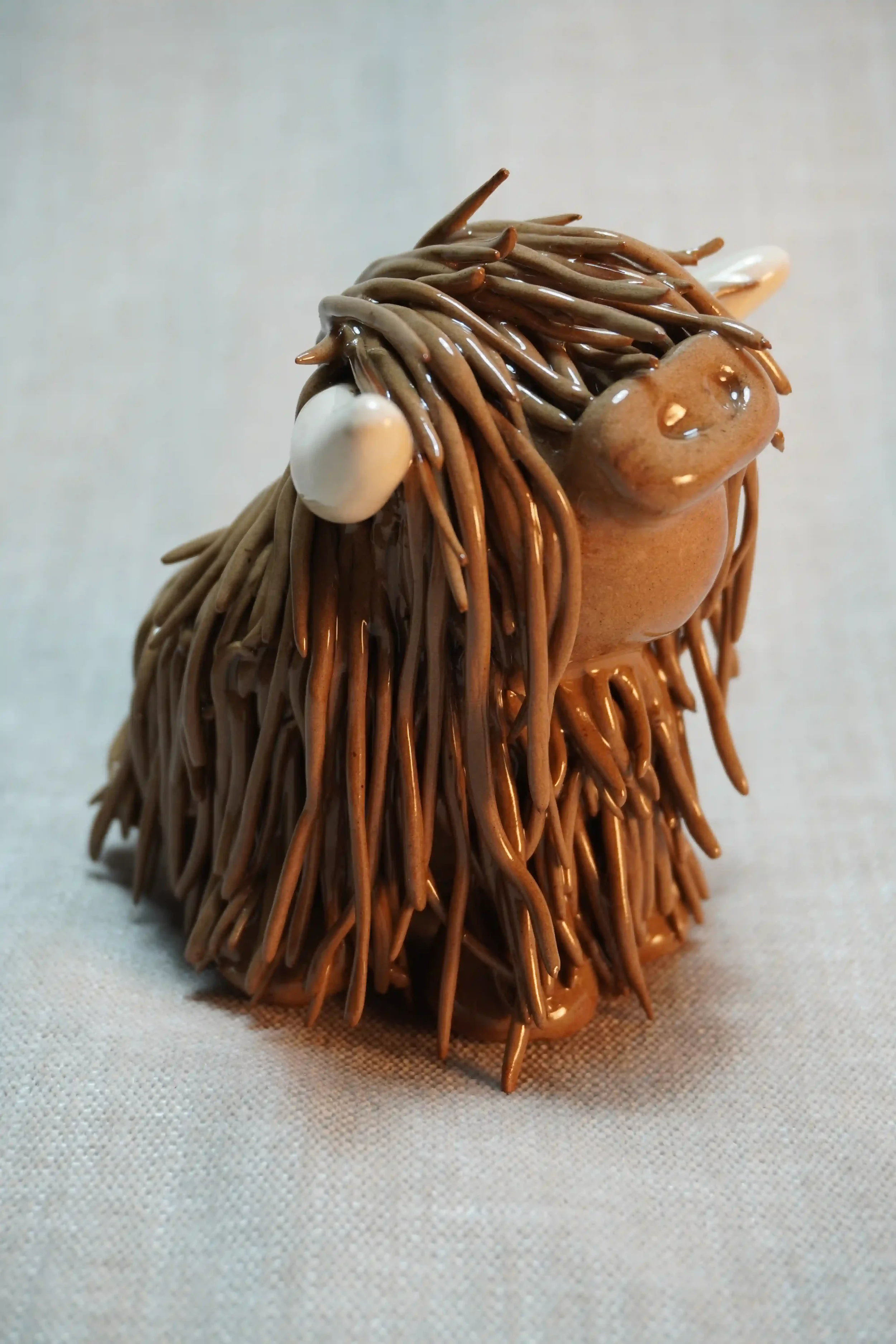 Side view of a medium brown handmade ceramic ornament of a Highland Cow with beige horns.
