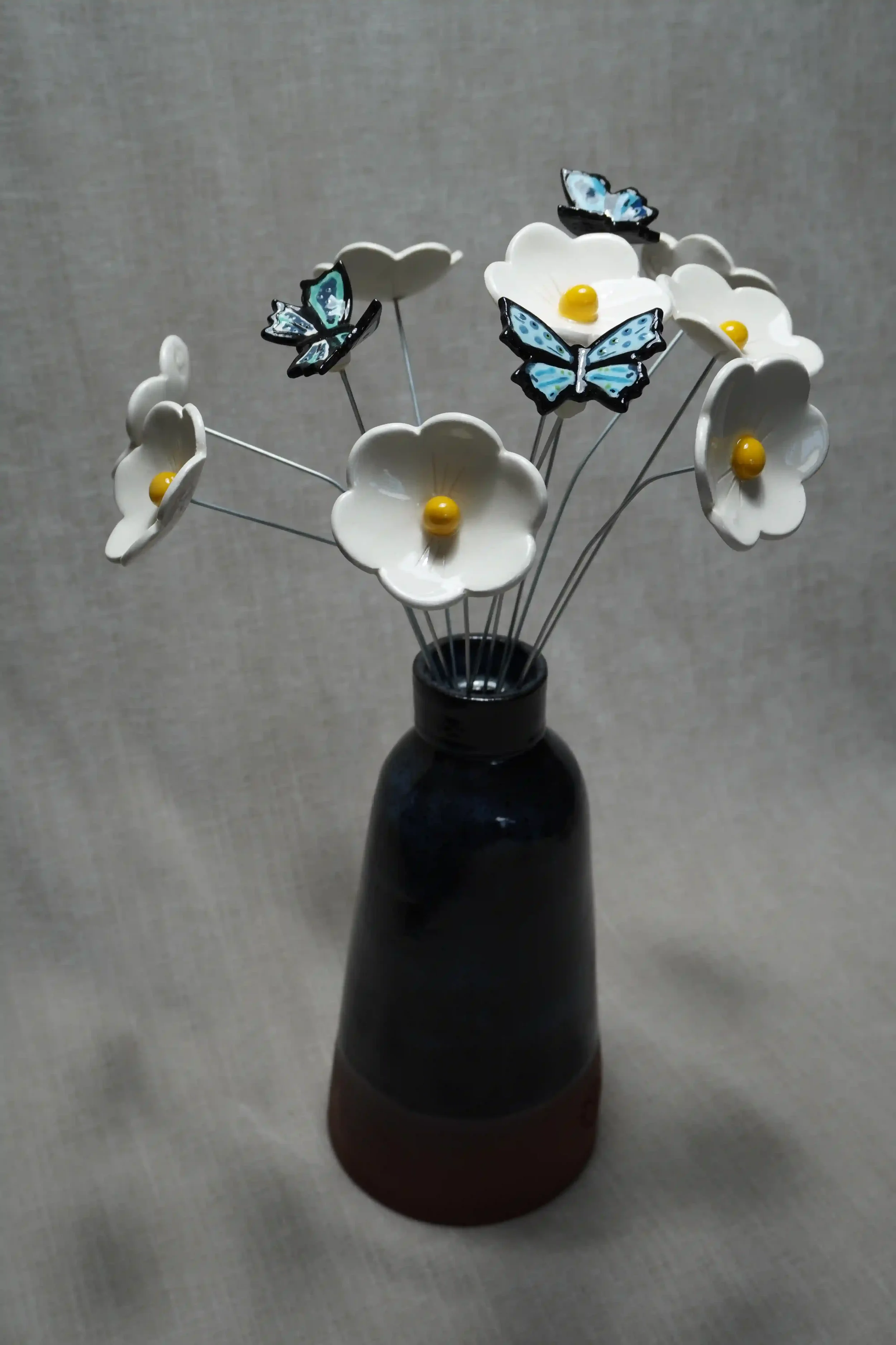 A blue vase with ceramic white flowers with yellow centres and ceramic blue butterflies attached to stems.