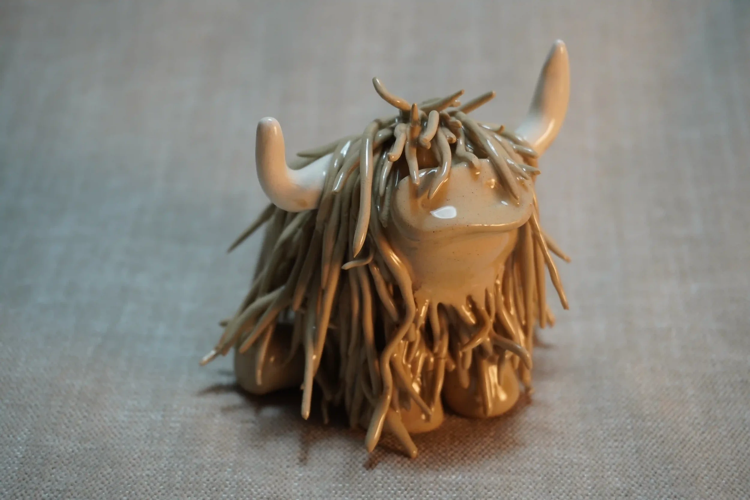 Front view of a ceramic sculpture of a Highland Cow ornament with light brown-coloured hair and curving beige horns.
