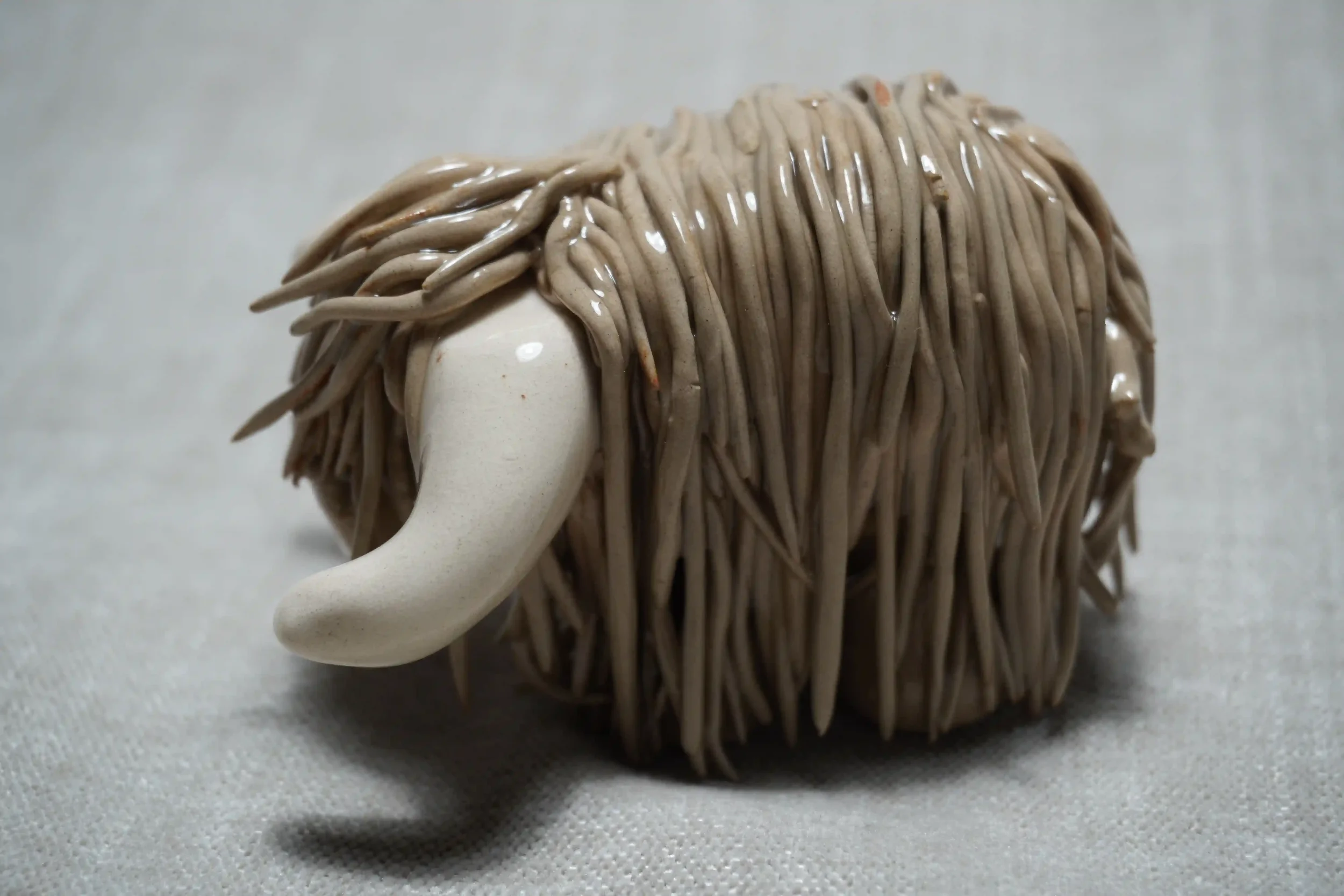 Side front view of a cute handmade ceramic Highland Cow ornament with light brown coloured hair and white horns.