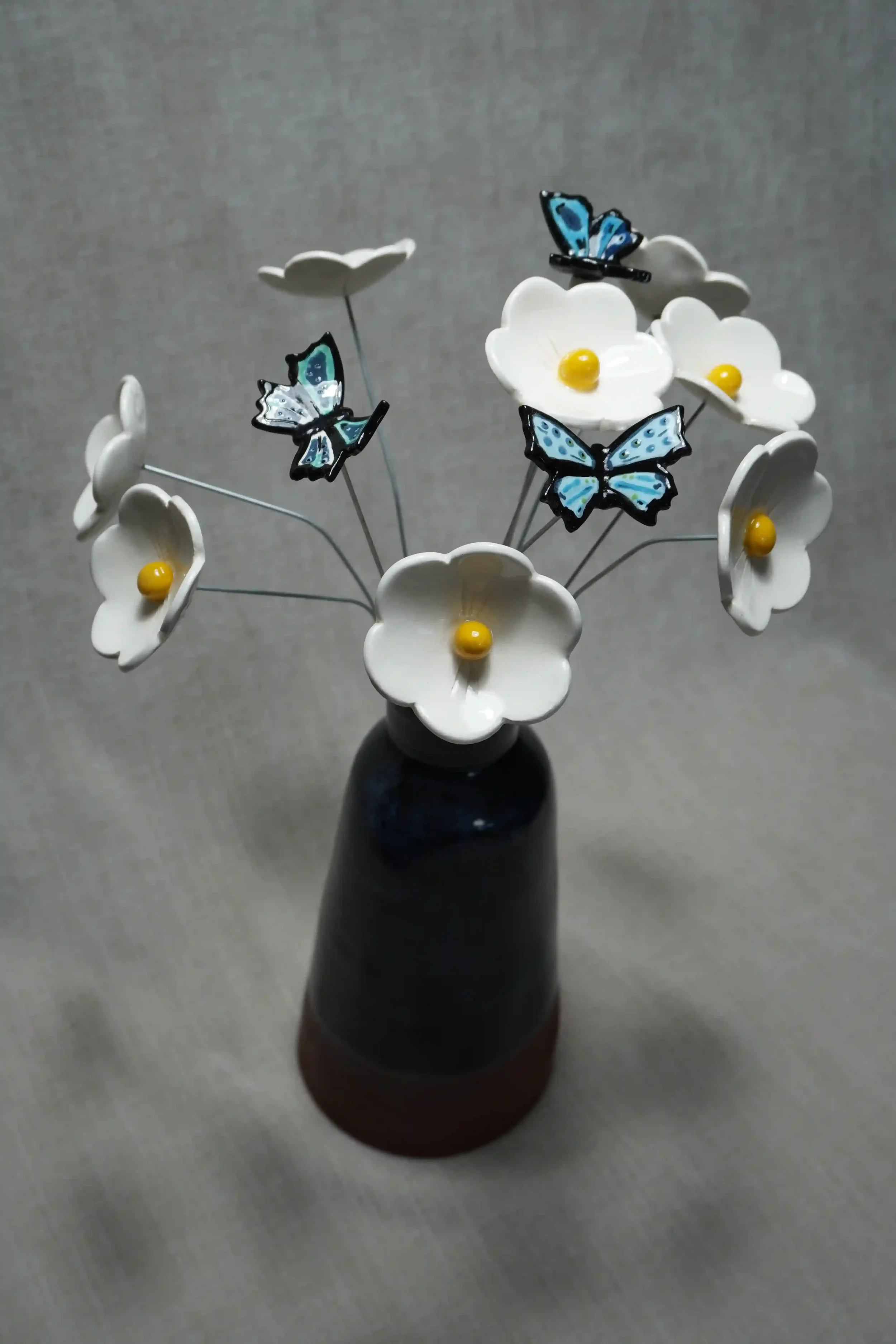 A dark blue-glazed handmade ceramic vase holding white ceramic Daisy flowers with yellow centers and three decorative ceramic blue butterflies, set against a neutral background.