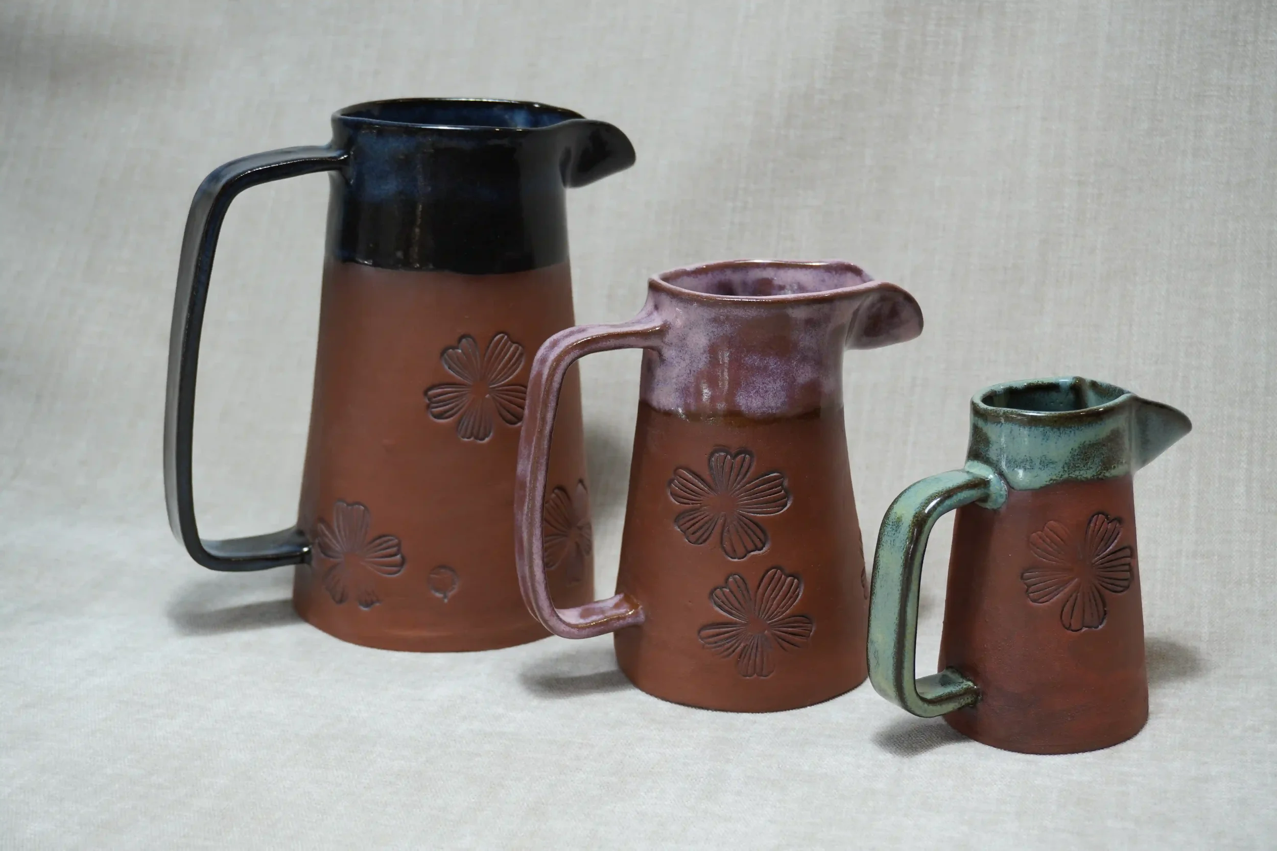 Three ceramic jugs (pitchers) with floral designs, varying in size and color, placed on a beige cloth background.