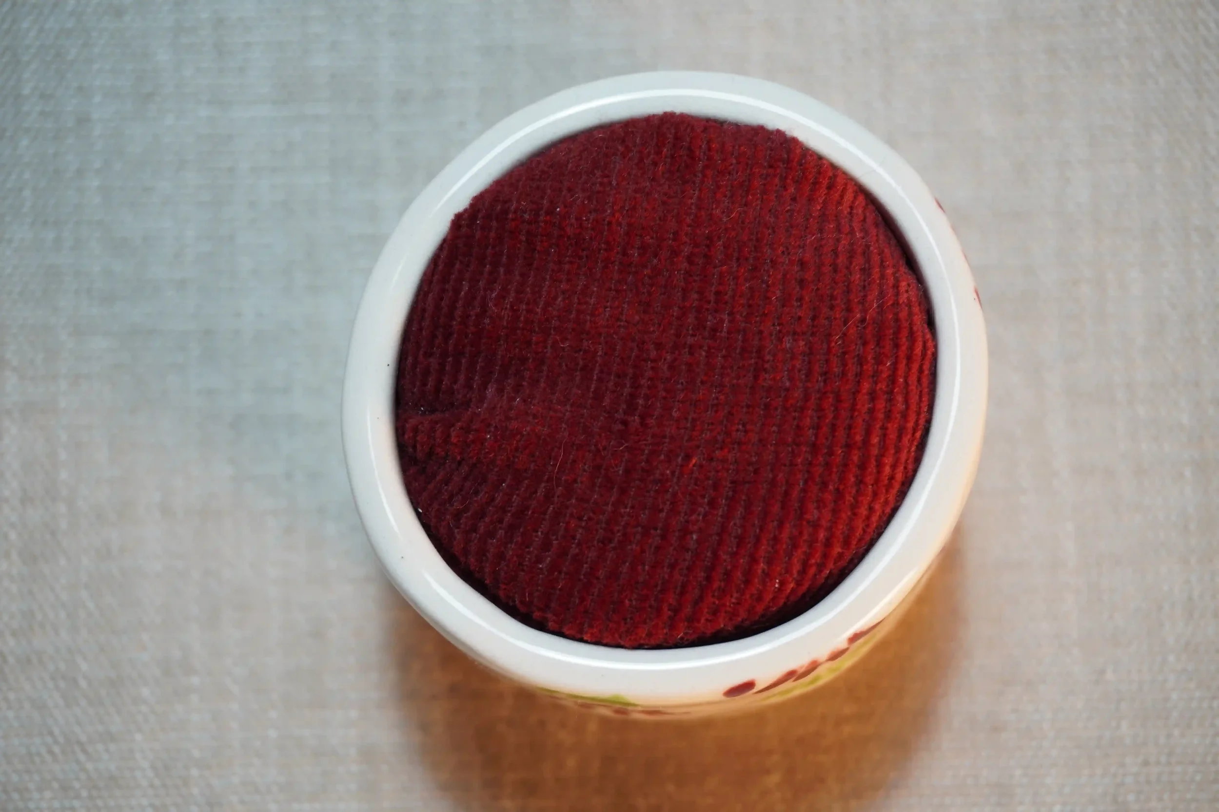 View from above of a ceramic pincushion with a red velvet cushion. The pincushion is shown on a textured surface.