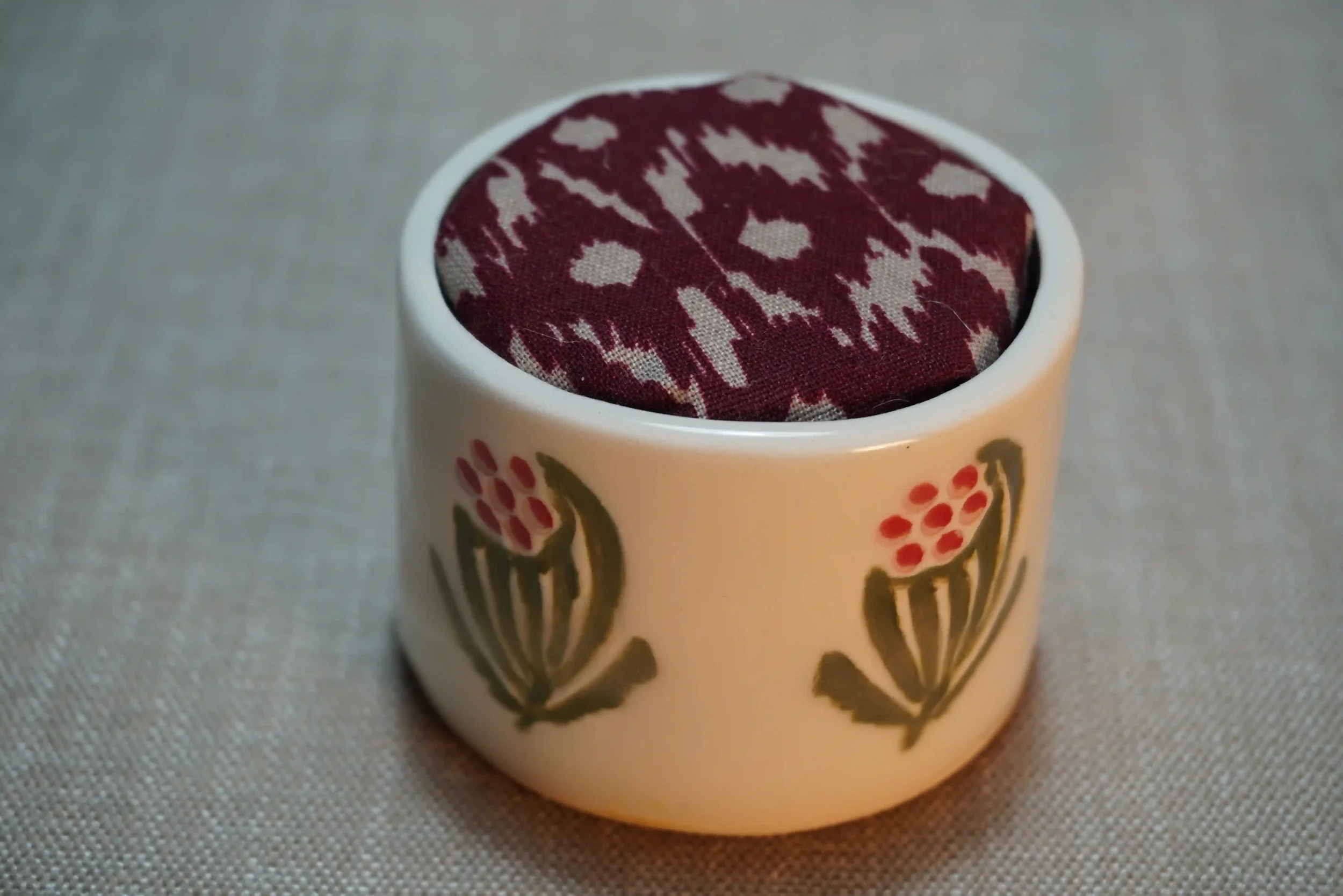 A ceramic pincushion with a floral design, filled with a maroon and white cushion. The pincushion is shown on a textured surface.