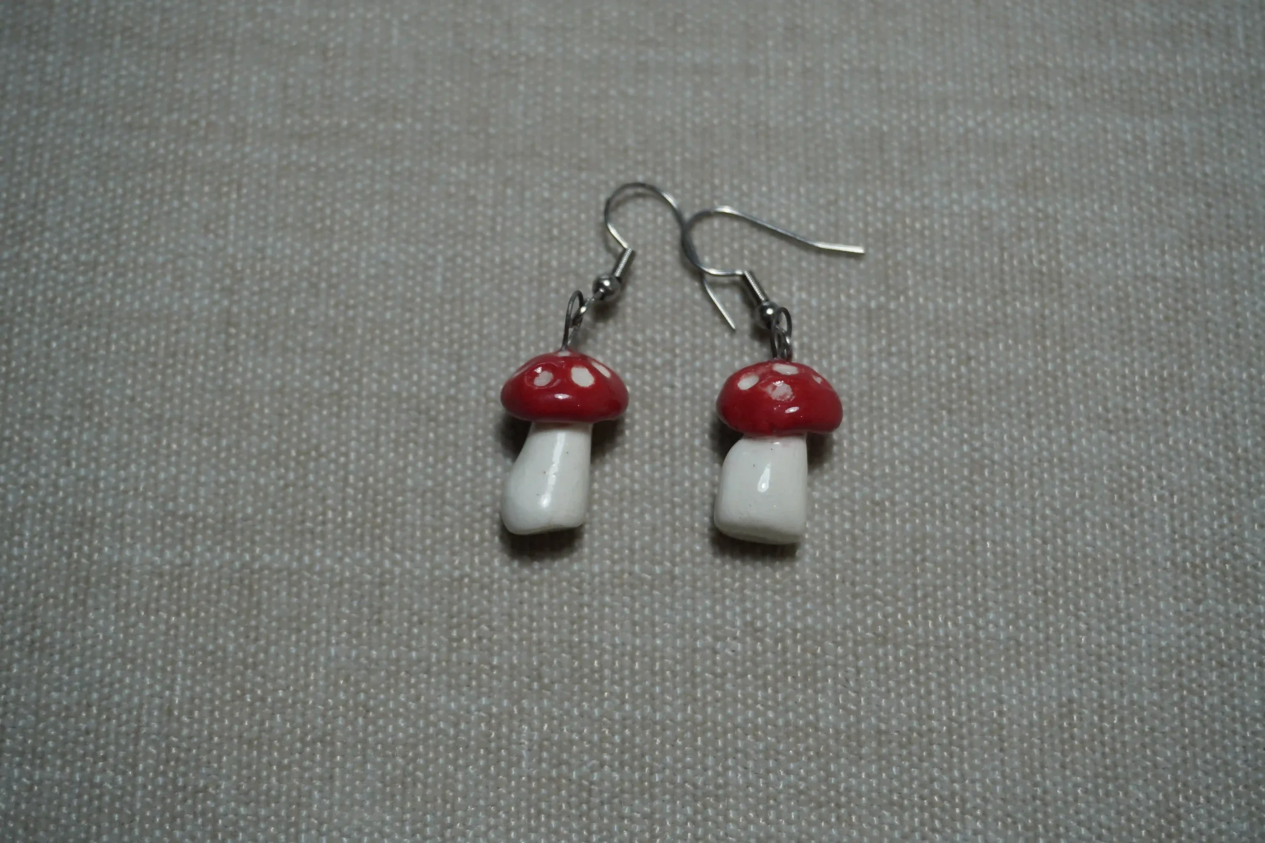 Pair of ceramic pottery mushroom-shaped earrings with red caps and white stems on a gray textured background.