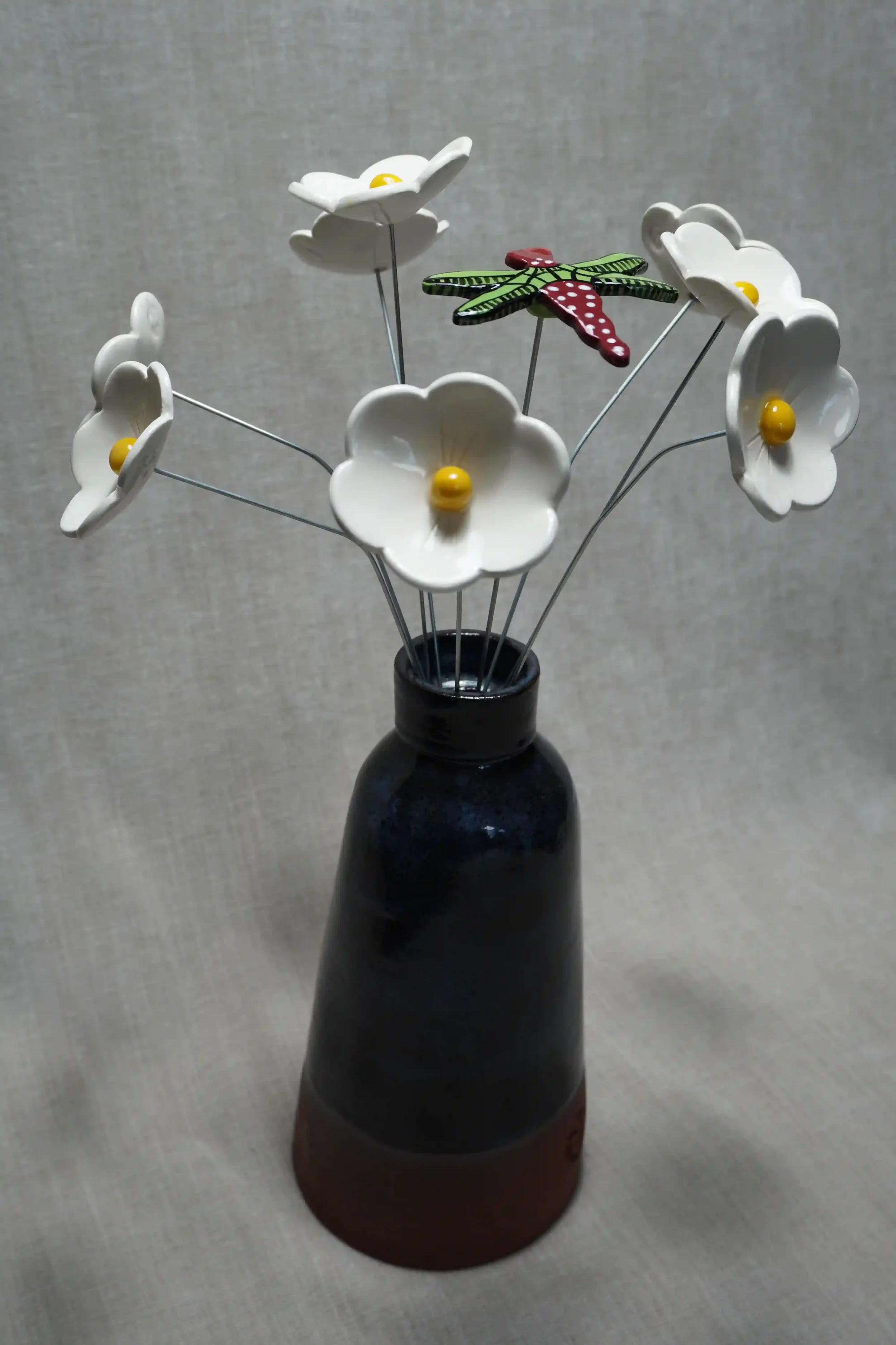 A dark blue and terracotta ceramic vase holding ceramic white Daisy flowers with yellow centers, and a colorful ceramic dragonfly with green and black wings and a red body with white speckles, on thin metal stems.