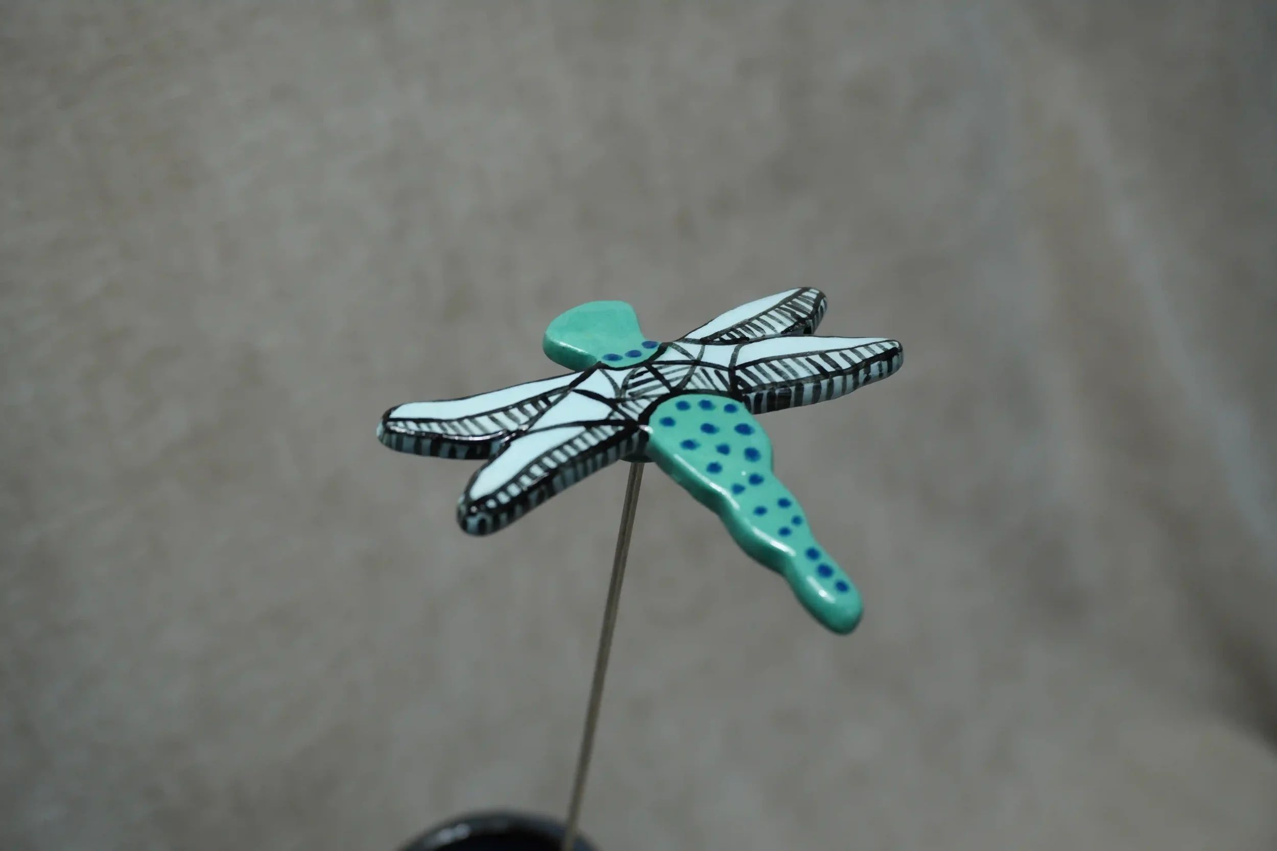 A ceramic pottery dragonfly with a teal body with blue specks and light blue and black wings on a metal stem.