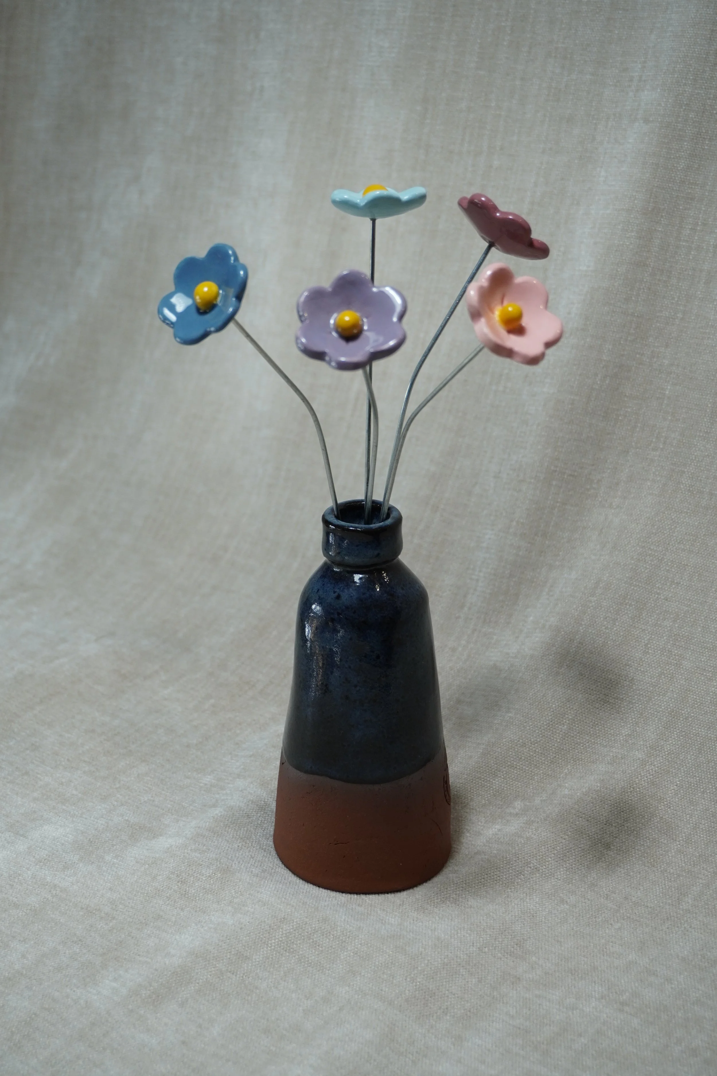 A ceramic pottery vase with a wide, rounded terracotta base, narrow neck, and a blue glaze, holding five ceramic purple, pink, light blue, and medium blue flowers with metal stems, displayed on a light neutral background.