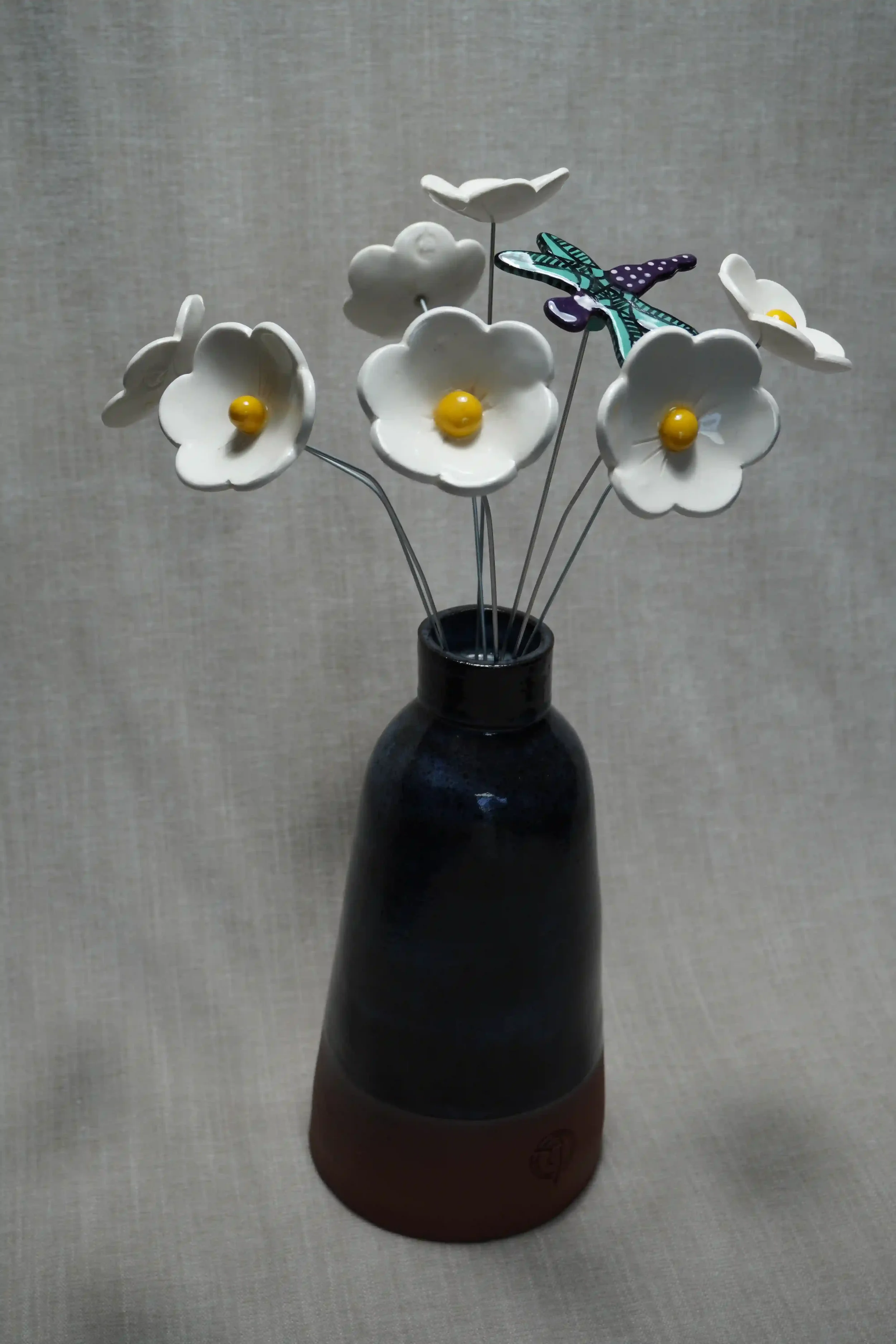 A dark blue and terracotta ceramic vase holding ceramic white Daisy flowers with yellow centers, a purple and teal dragonfly with black spots, on thin metal stems.