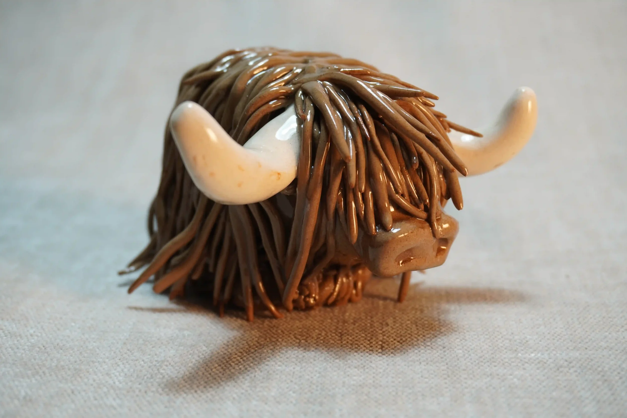 Quarter front view of a handmade pottery ceramic sculpture of a Highland Cow ornament with milk chocolate-coloured hair and beige horns.