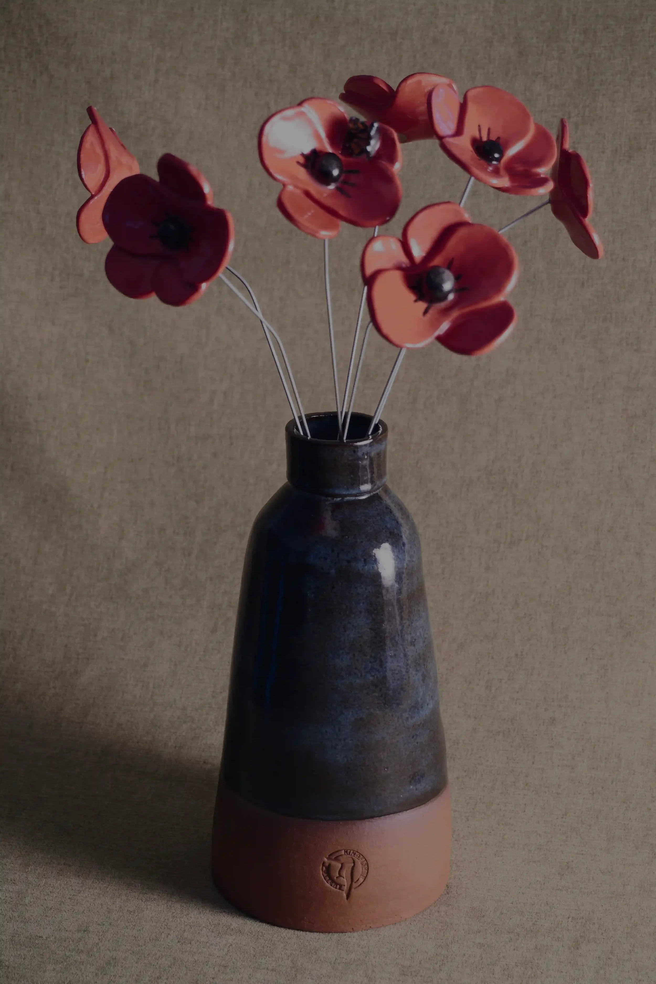 A ceramic pottery vase with a wide, rounded terracotta base, narrow neck, and a blue glaze, holding seven red ceramic poppies with black centres and metal stems on a light brown background.
