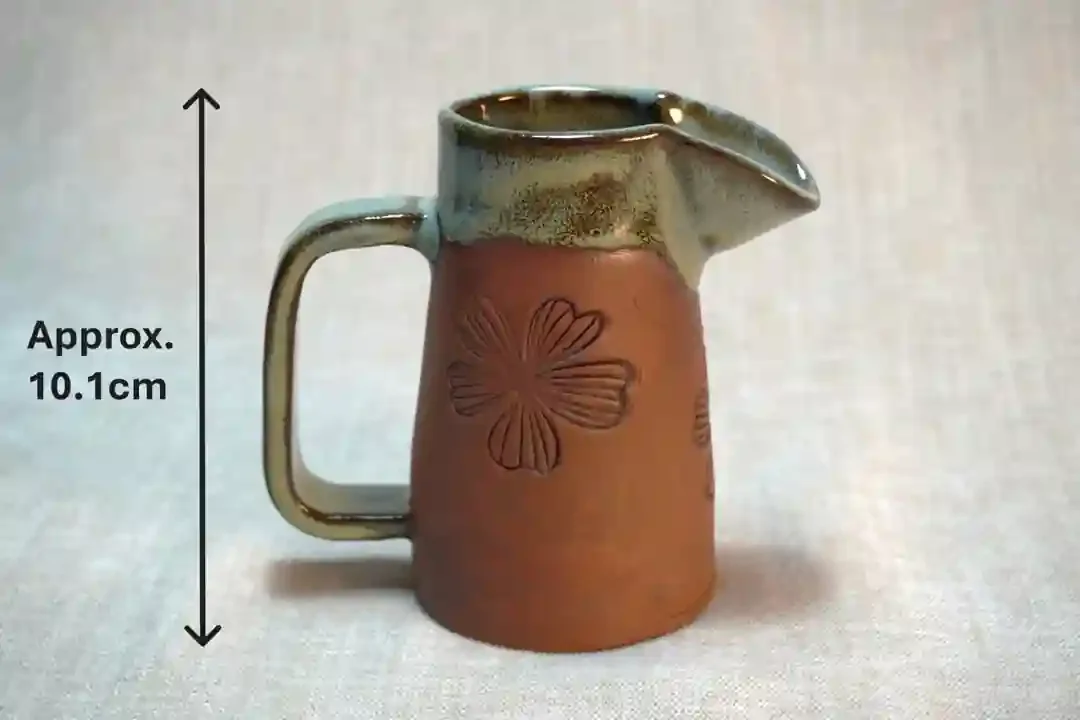 A small ceramic pitcher with embossed floral design on the terracotta-colored body, glazed in light green, with a handle on the side and a spout on top. Approximately 10.1 centimeters tall.