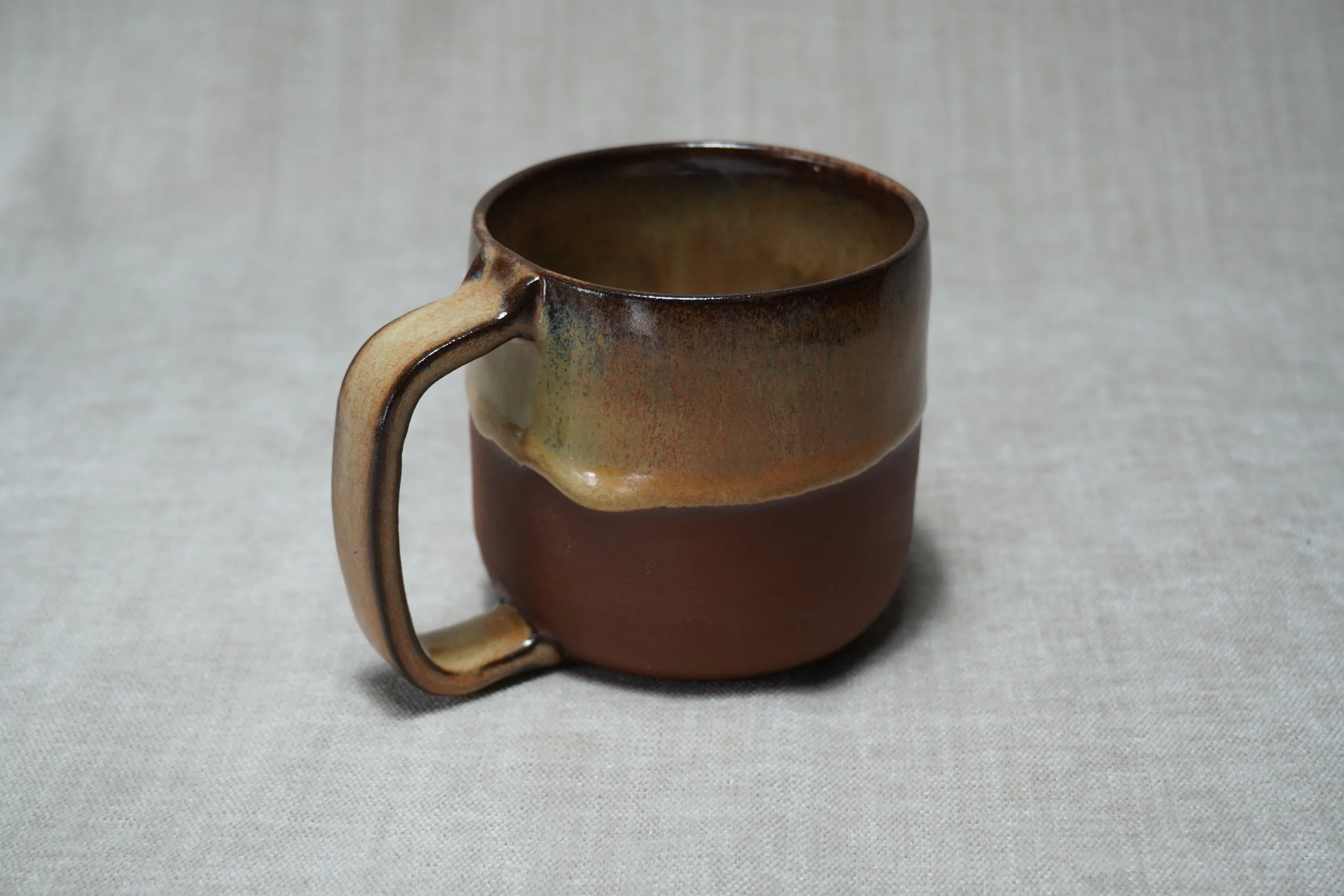 A ceramic mug with a gradient glaze from dark brown to light honey colour on a light, textured fabric background.