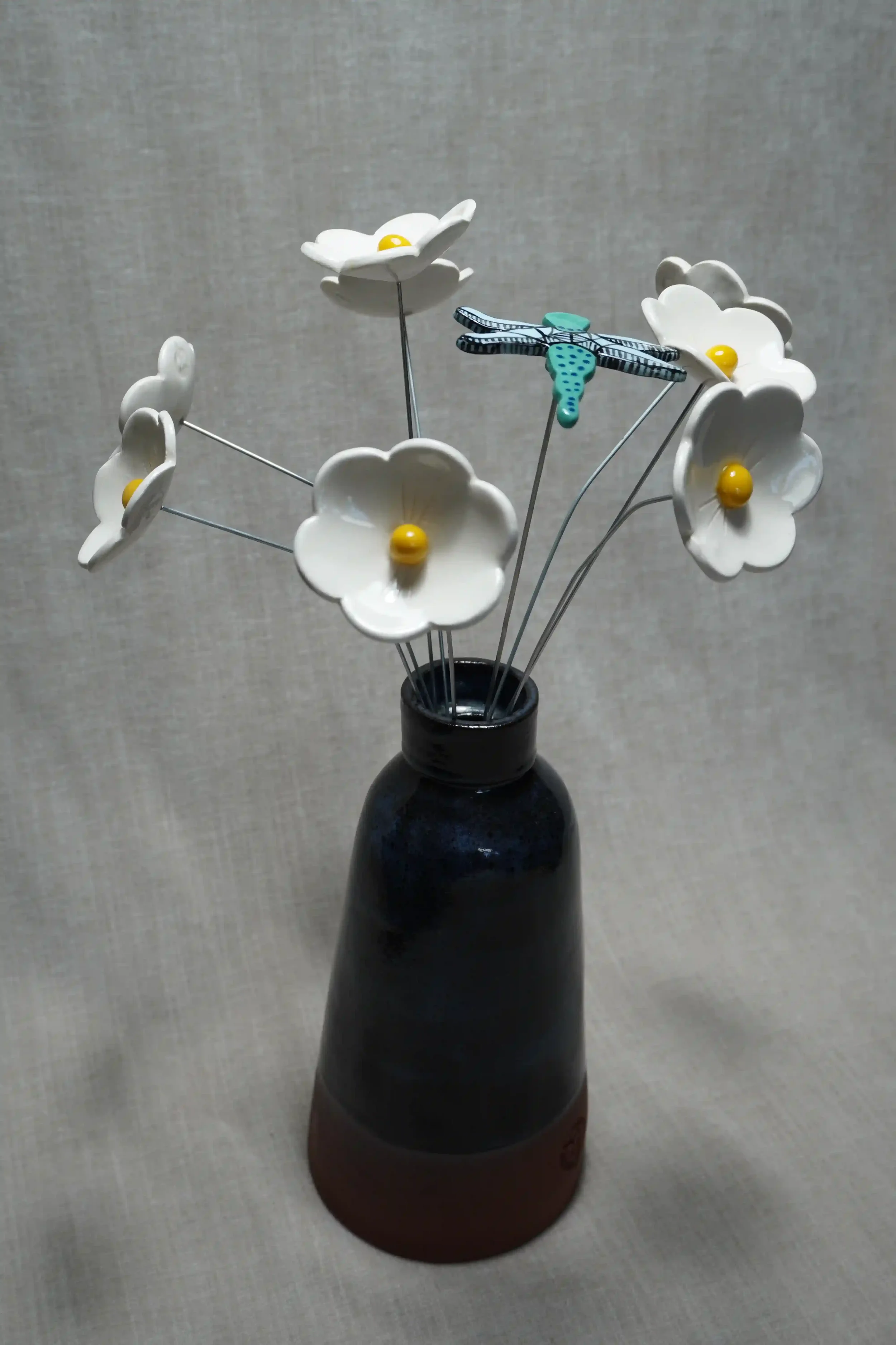 A dark blue and terracotta ceramic vase holding ceramic white Daisy flowers with yellow centers, a dragonfly with turquoise body with blue spots, black and light blue wings, on thin metal stems.  