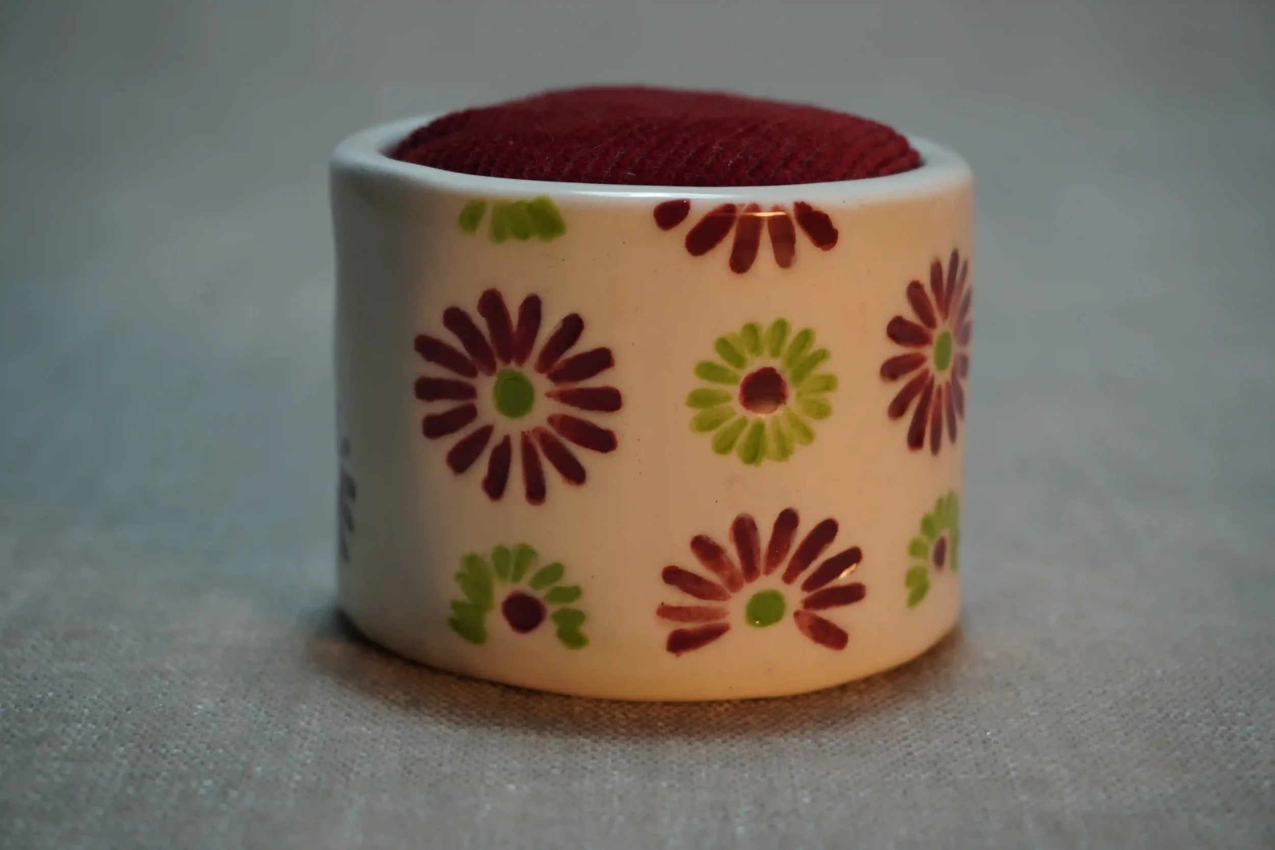 A ceramic pincushion with a red and green firework pattern , filled with red velvet cushion. The pincushion is shown on a textured surface.