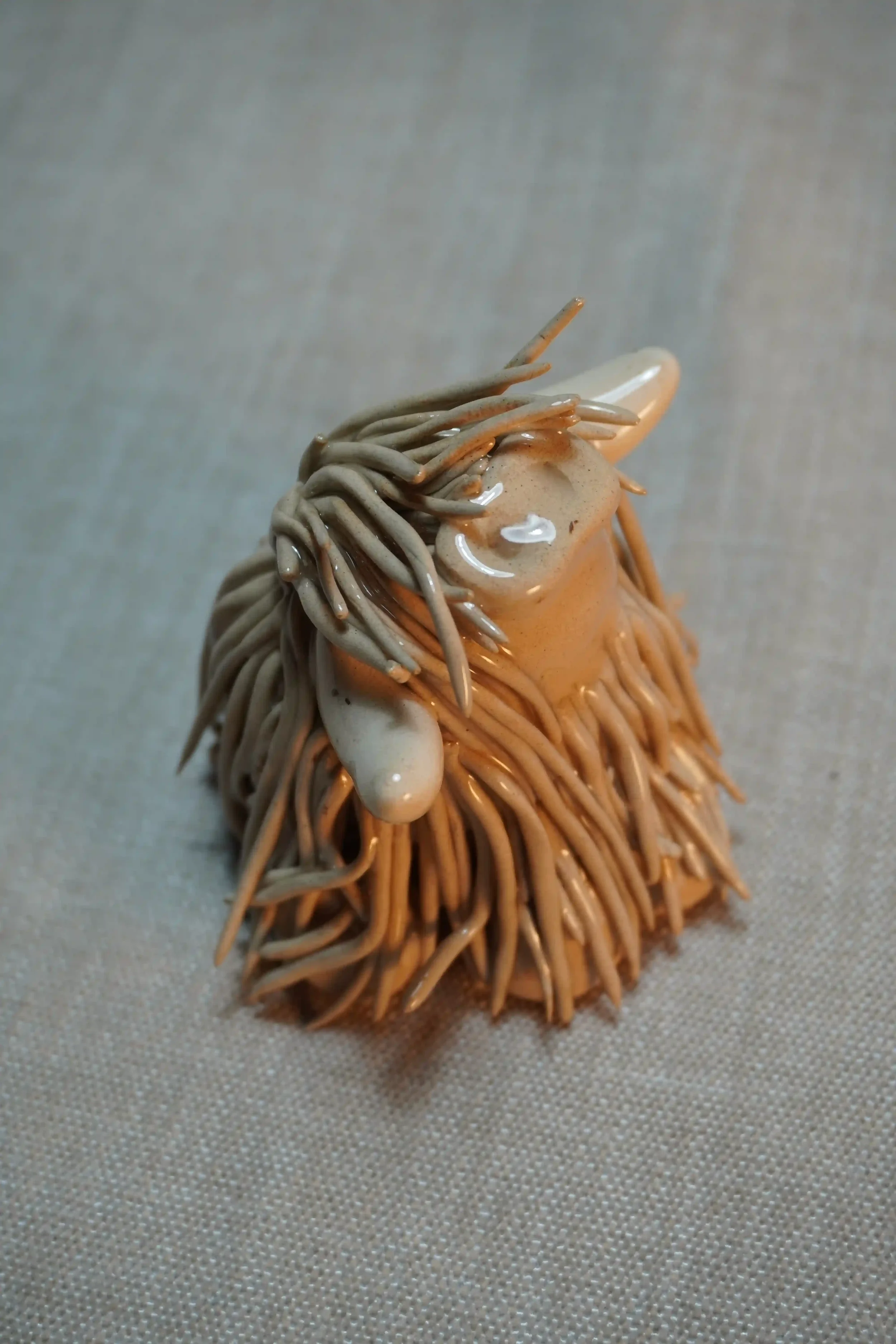 Side view of a handmade ceramic sculpture of a Highland Cow ornament with light brown-coloured hair and beige horns.