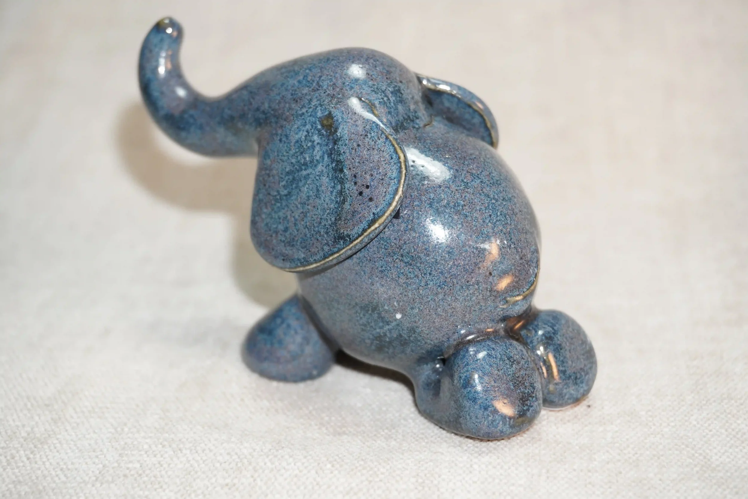 	
A handmade small ceramic elephant figurine with a speckled blue glossy glaze finish and it's trunk in the air.