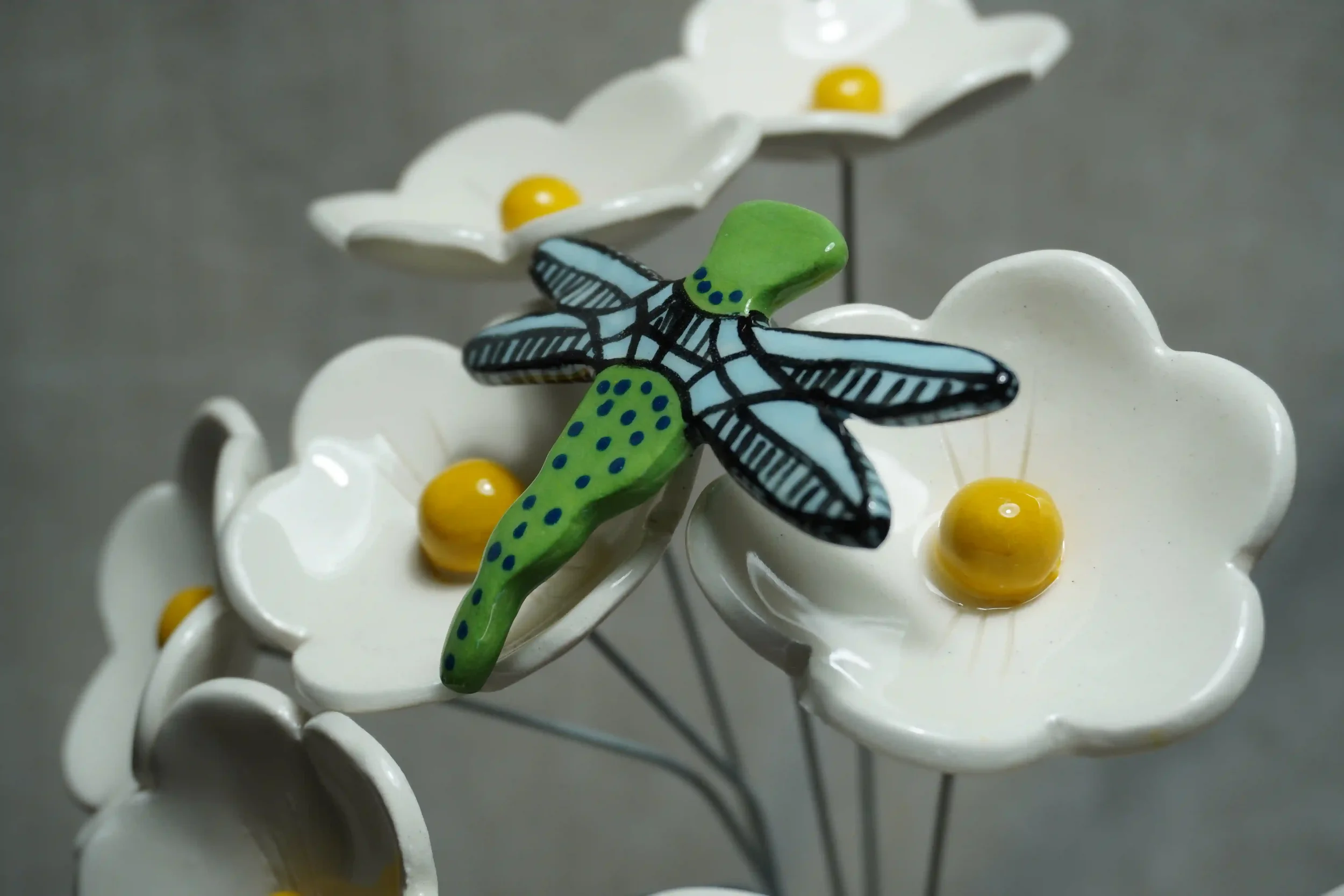 A close-up of a colorful ceramic dragonfly with light blue wings and a green body with blue speckles, set amongst white ceramic daisies with yellow centres. All mounted on thin metal stems, against a plain gray background