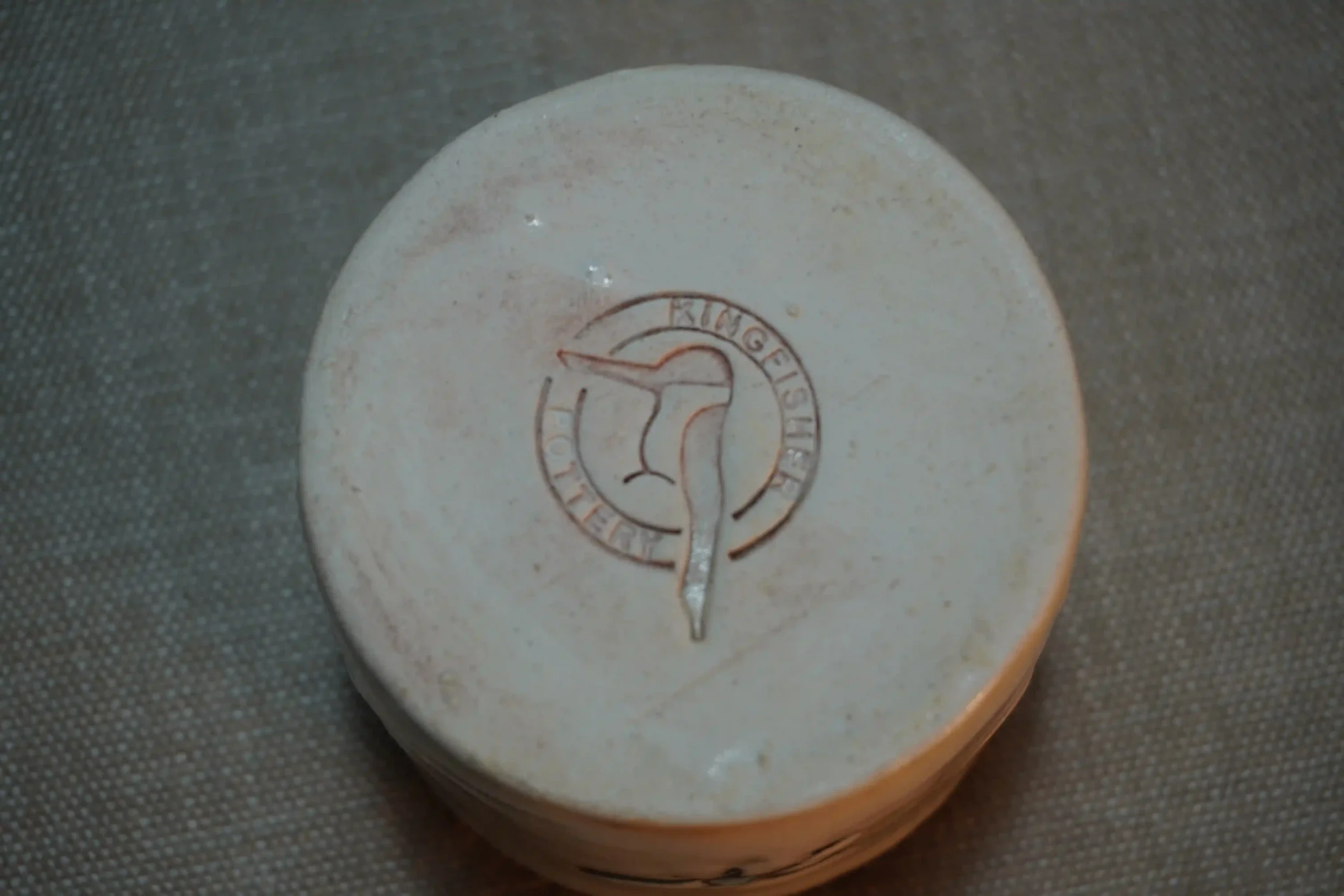 View of a pottery maker's mark on the bottom of a ceramic pincushion. The maker's mark shows a kingfisher and words saying Kingfisher Pottery.