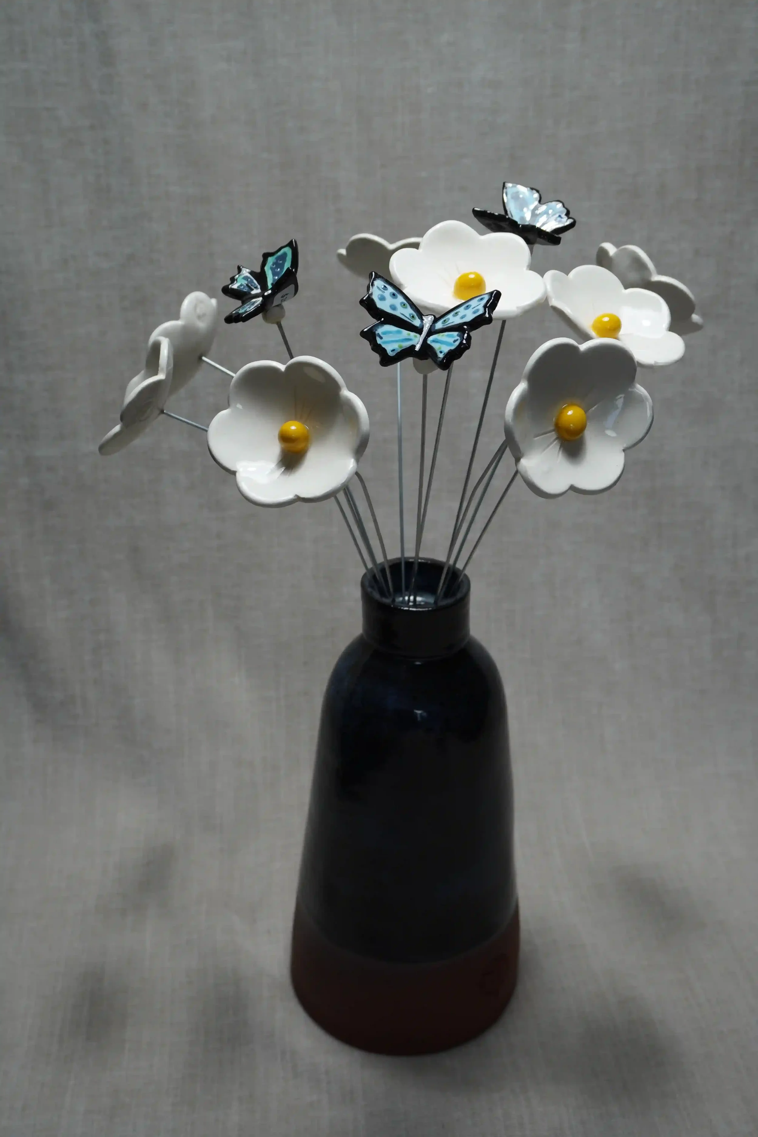 A dark blue-glazed handmade ceramic vase holding white ceramic Daisy flowers with yellow centers and three decorative ceramic blue butterflies, set against a neutral background.