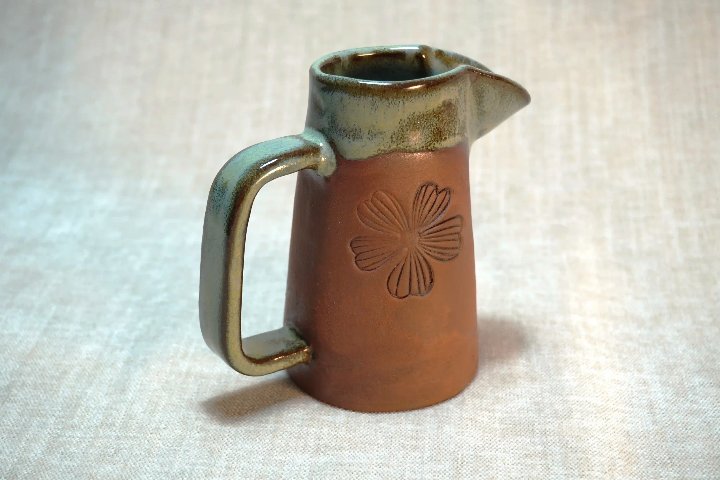 A small ceramic pitcher with embossed floral design on the terracotta-colored body, glazed in light green, with a handle on the side and a spout on top.