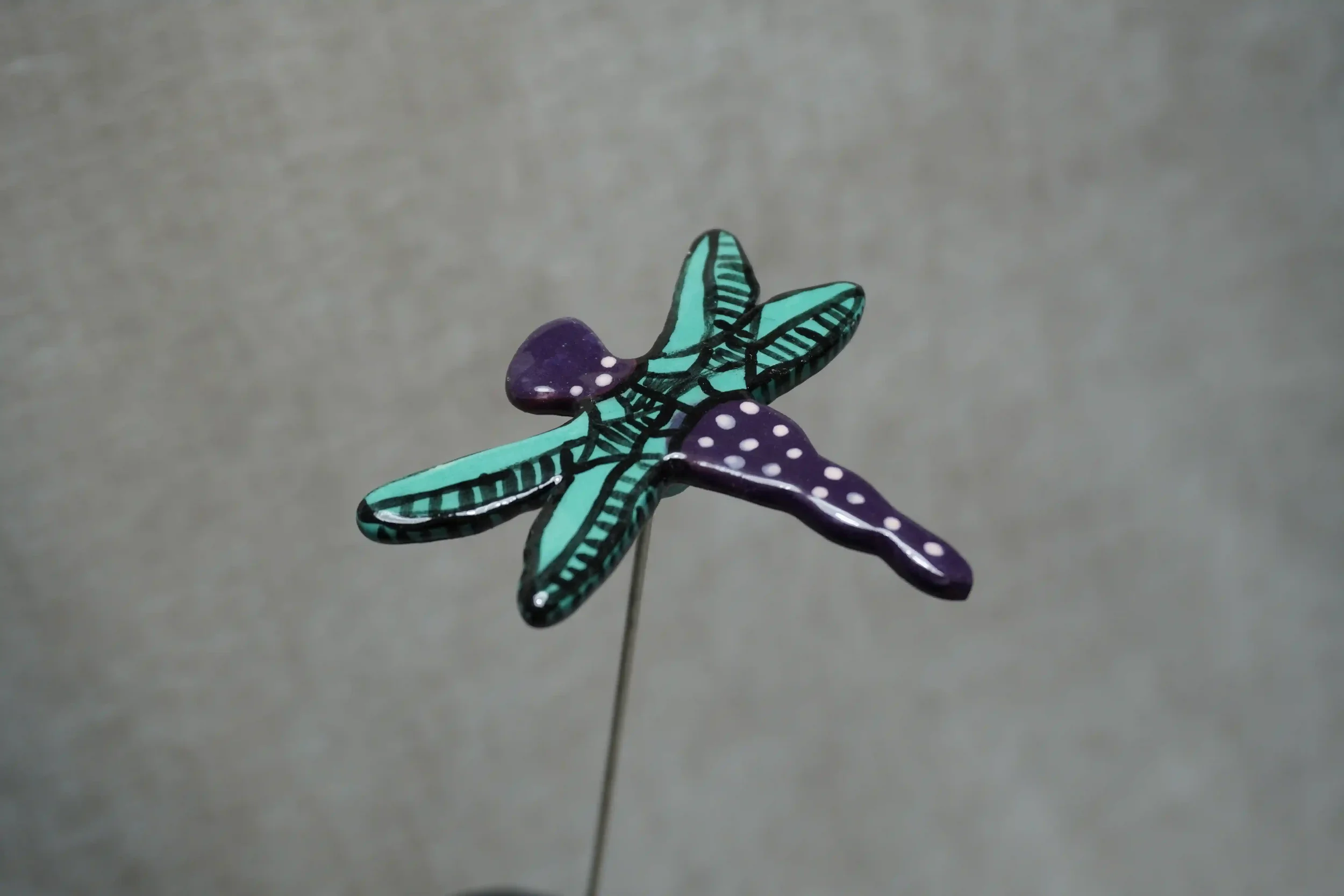 Colorful ceramic dragonfly with teal and black wings and a purple body with white speckles, mounted on a thin metal stem, against a plain gray background.