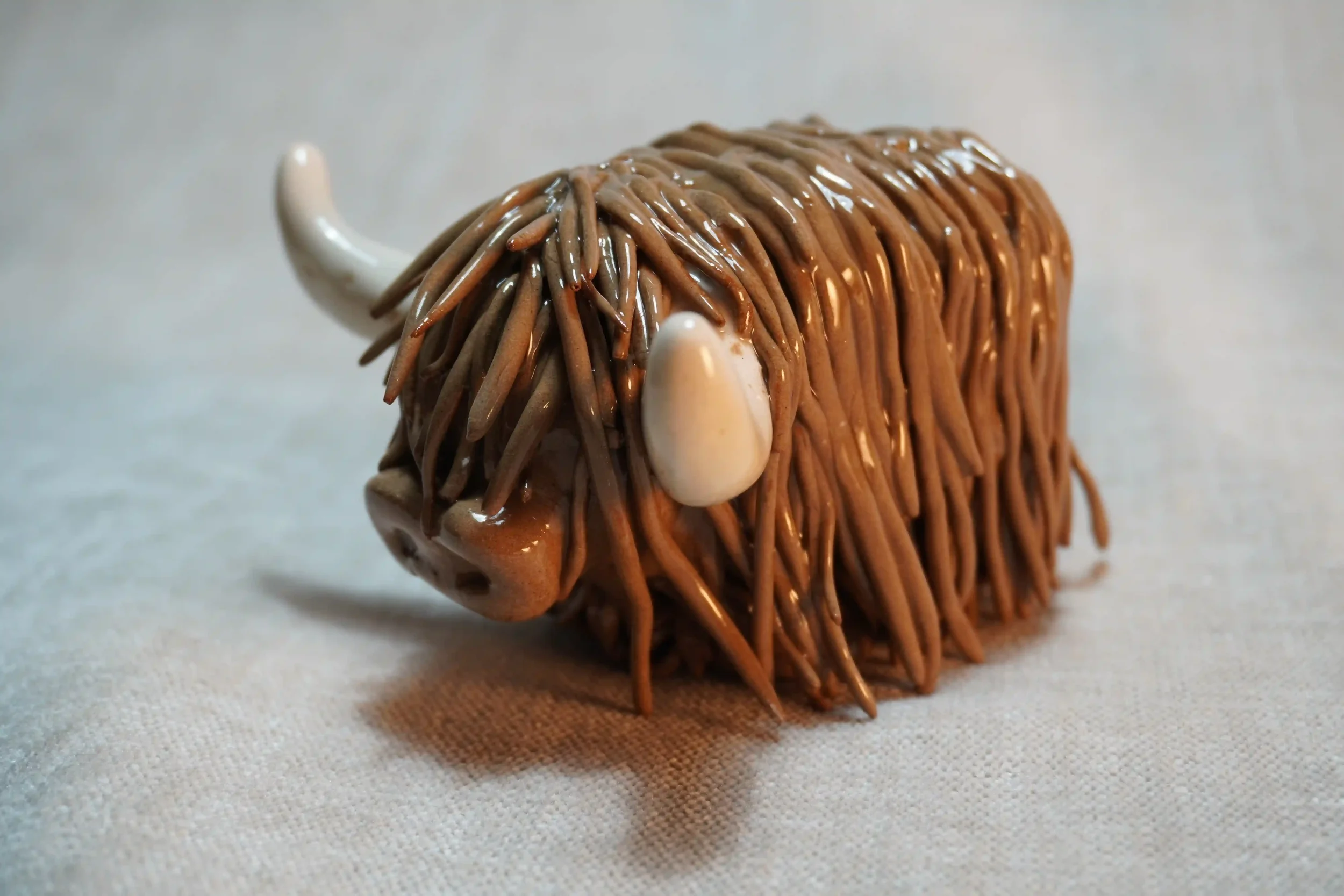 Quarter front view of a handmade pottery ceramic sculpture of a Highland Cow ornament with milk chocolate-coloured hair and beige horns.