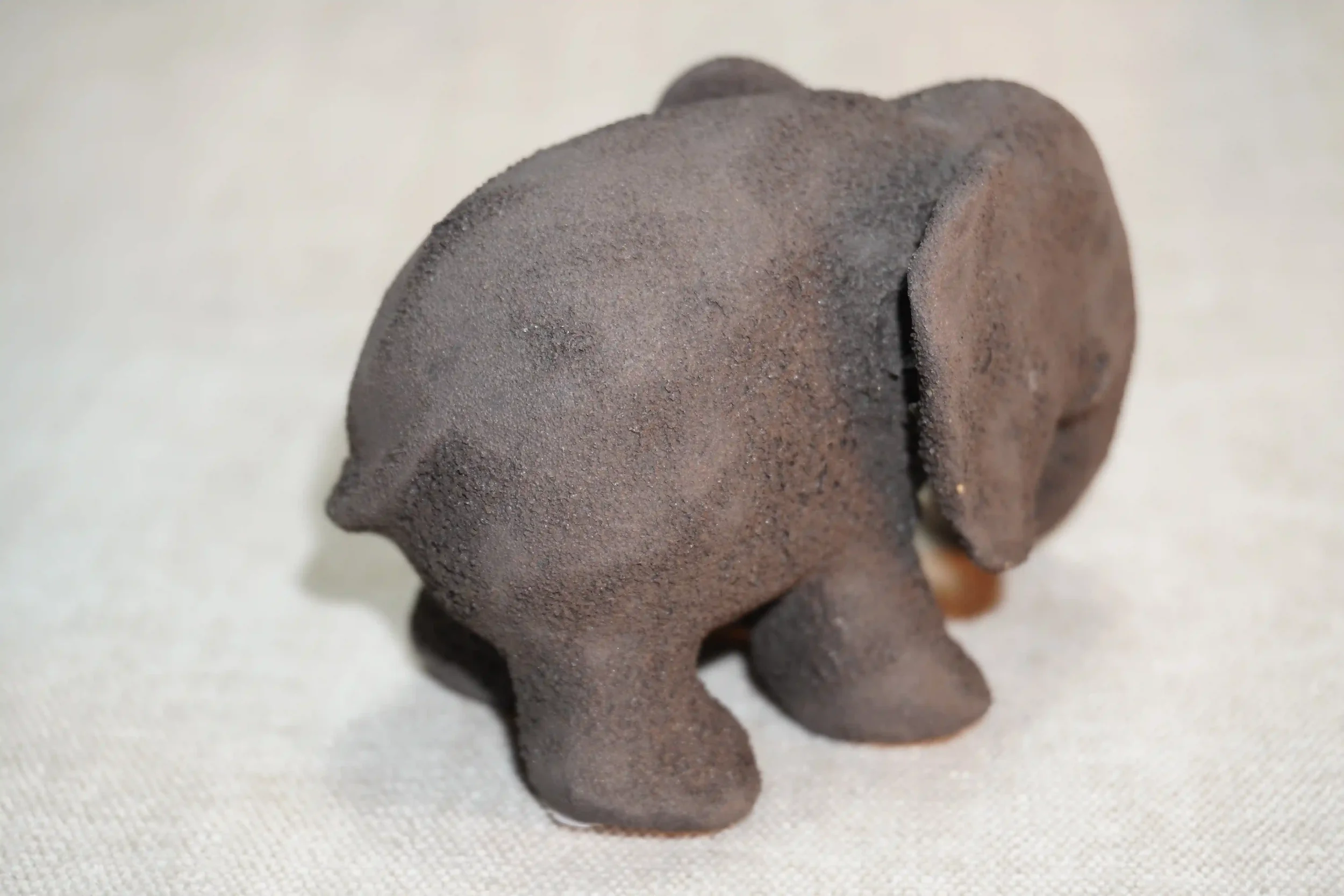 Rear view of a handmade pottery elephant figurine with a dark grey textured surface and holding a branch in it's trunk, placed on a light-colored fabric surface.
