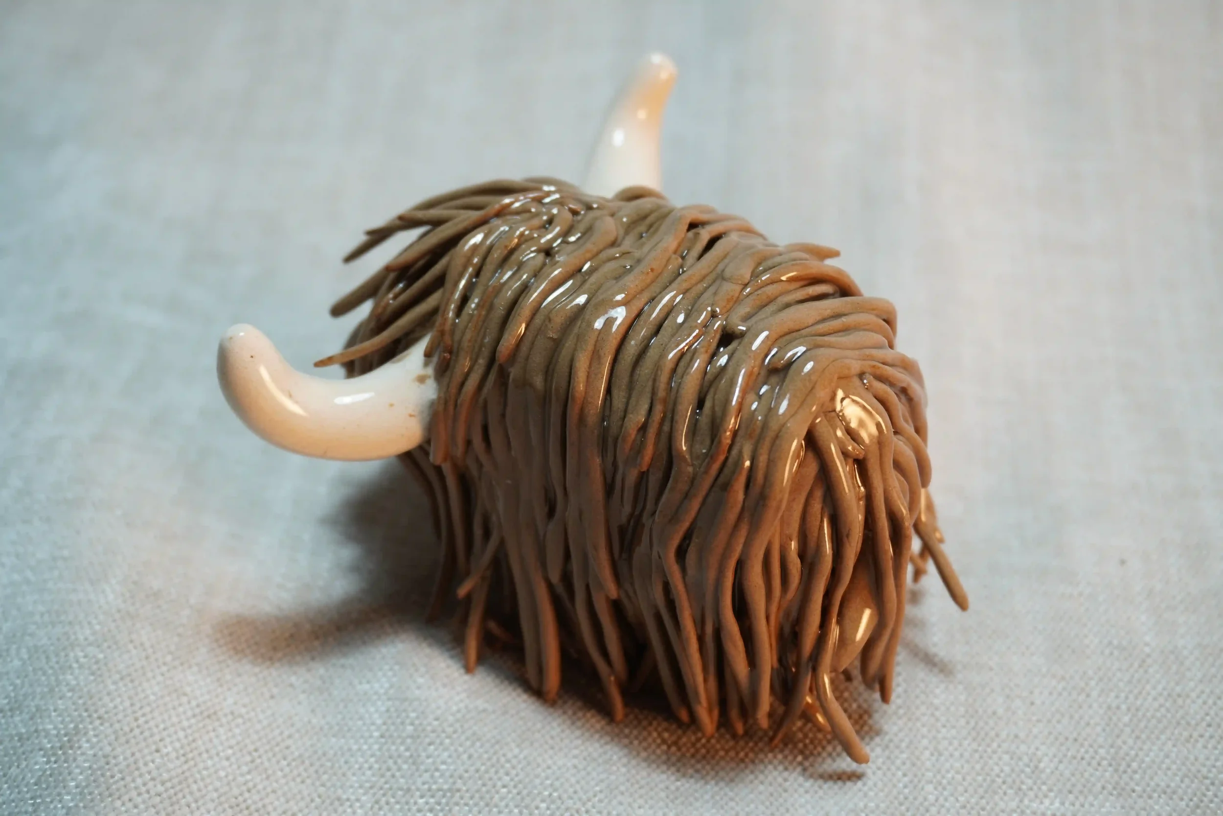 Quarter rear view of a handmade pottery ceramic sculpture of a Highland Cow ornament with milk chocolate-coloured hair and beige horns.