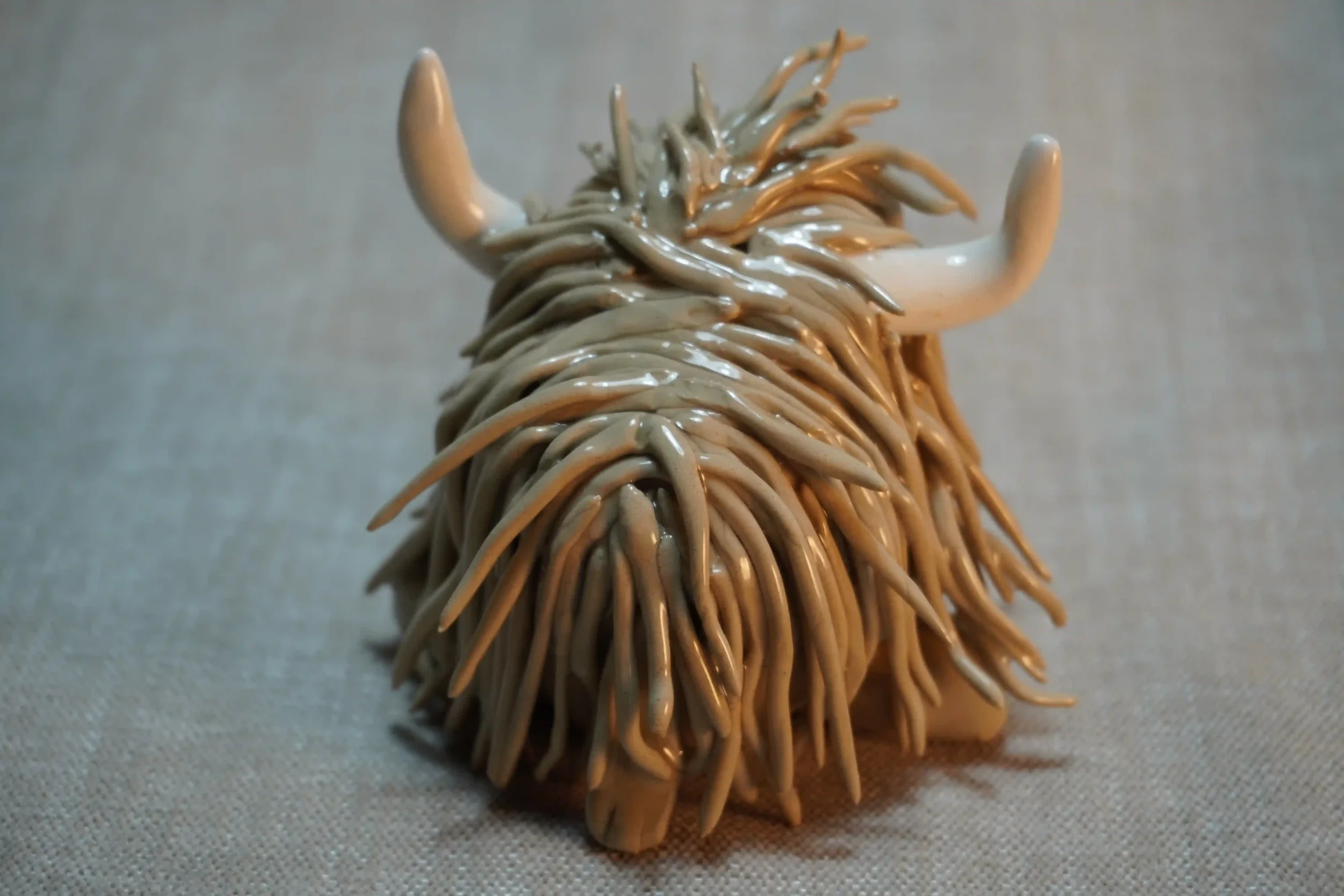 Rear view of a ceramic sculpture of a Highland Cow ornament with light brown-coloured hair and curving beige horns.
