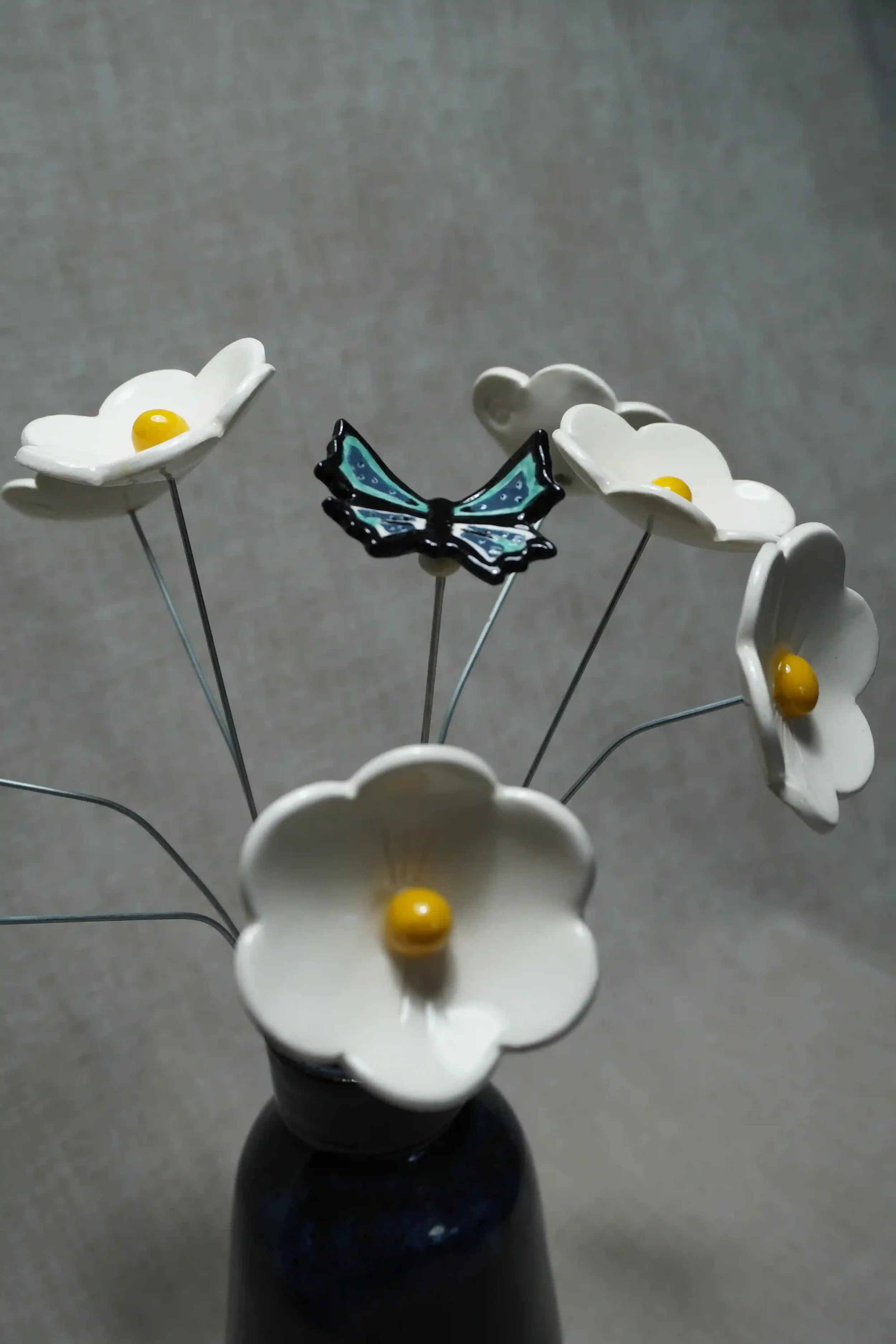 A decorative flower arrangement with ceramic white Daisy flowers featuring yellow centers, a blue and black ceramic butterfly, all on thin metal stems in a dark vase against a gray background.