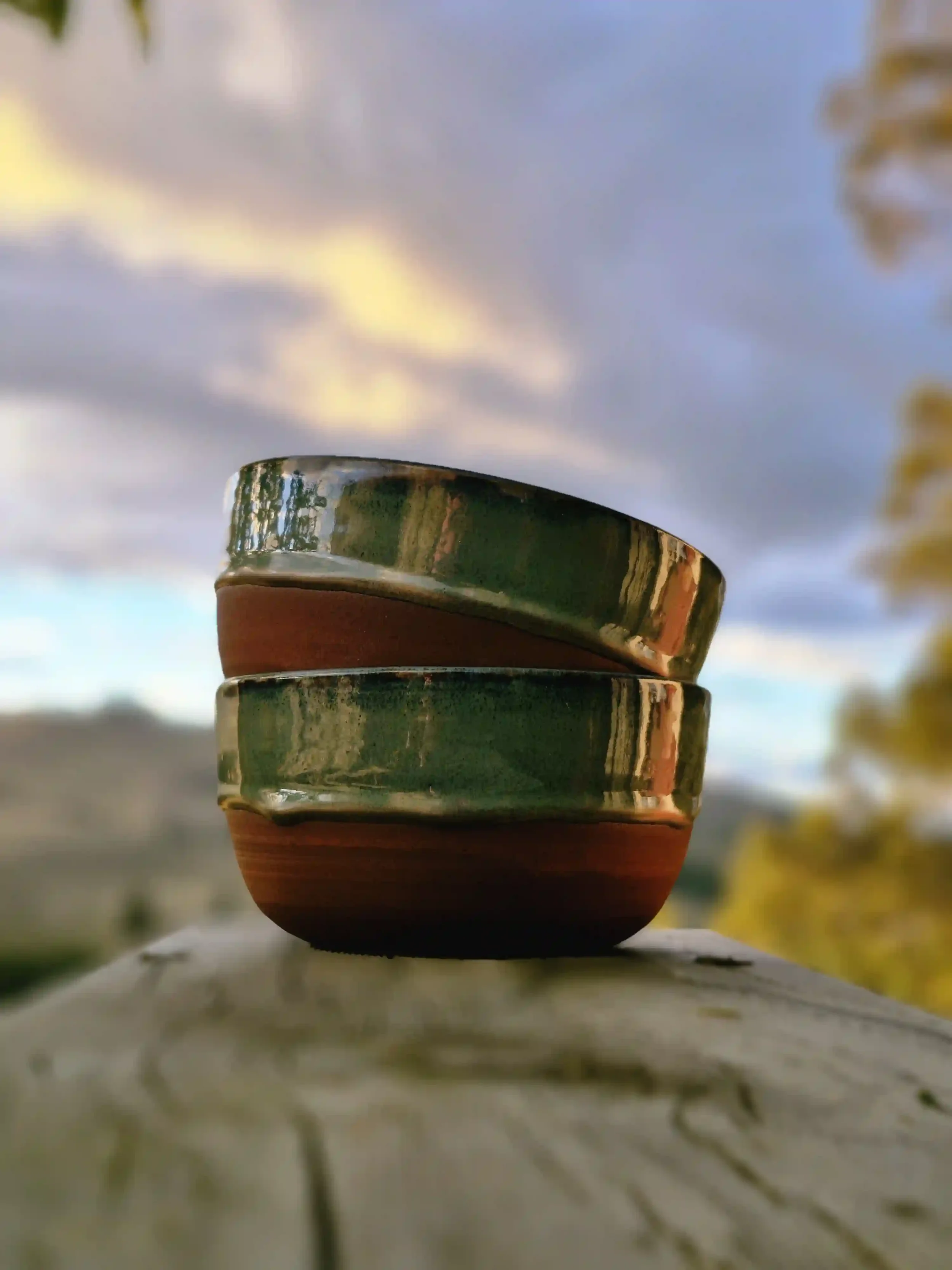 Two ceramic bowls stacked on a rustic wooden surface outdoors with a blurred landscape and sky in the background.