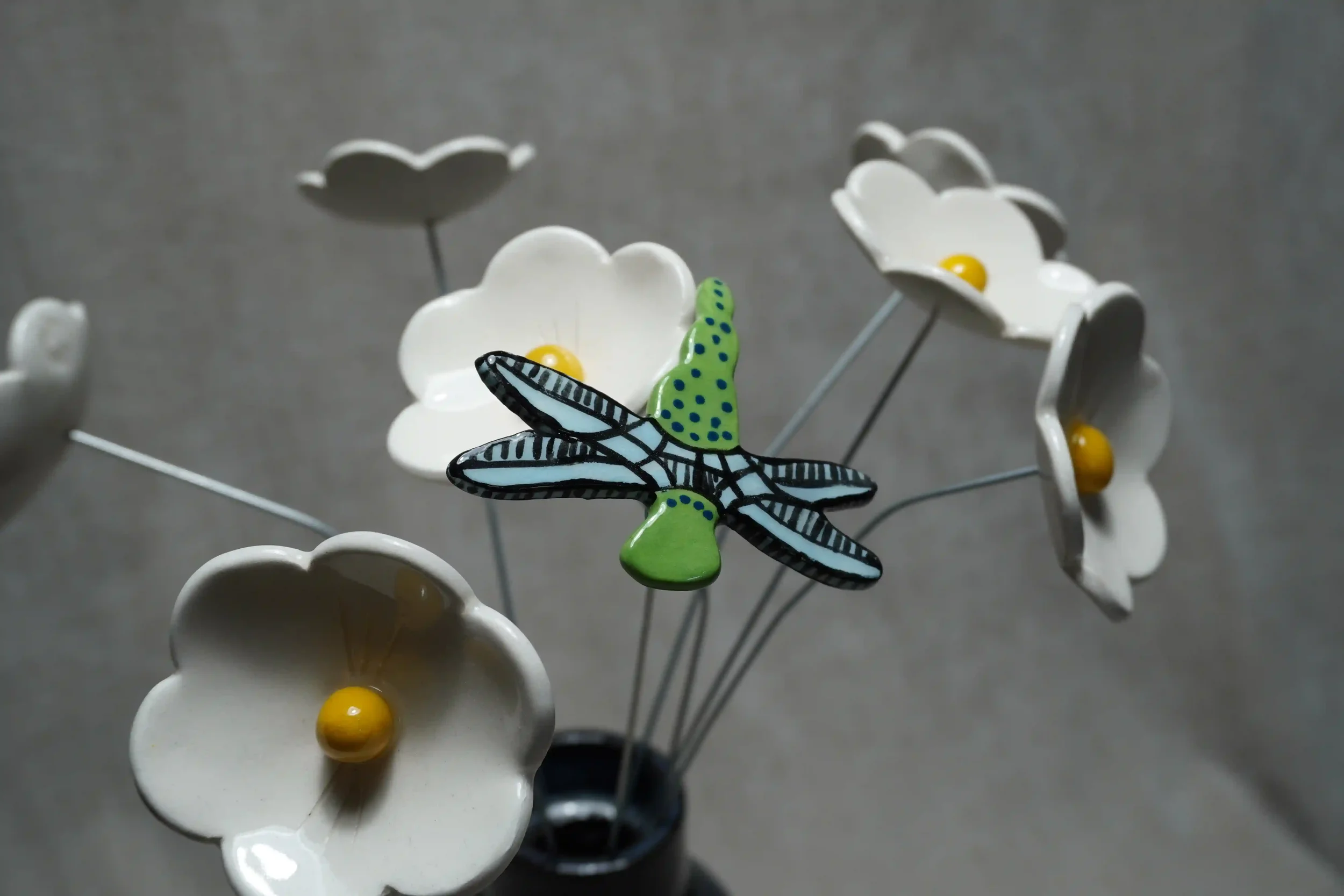 	
A close-up of a colorful ceramic dragonfly with light blue wings and a green body with blue speckles, set amongst white ceramic daisies with yellow centres. All mounted on thin metal stems, against a plain gray background