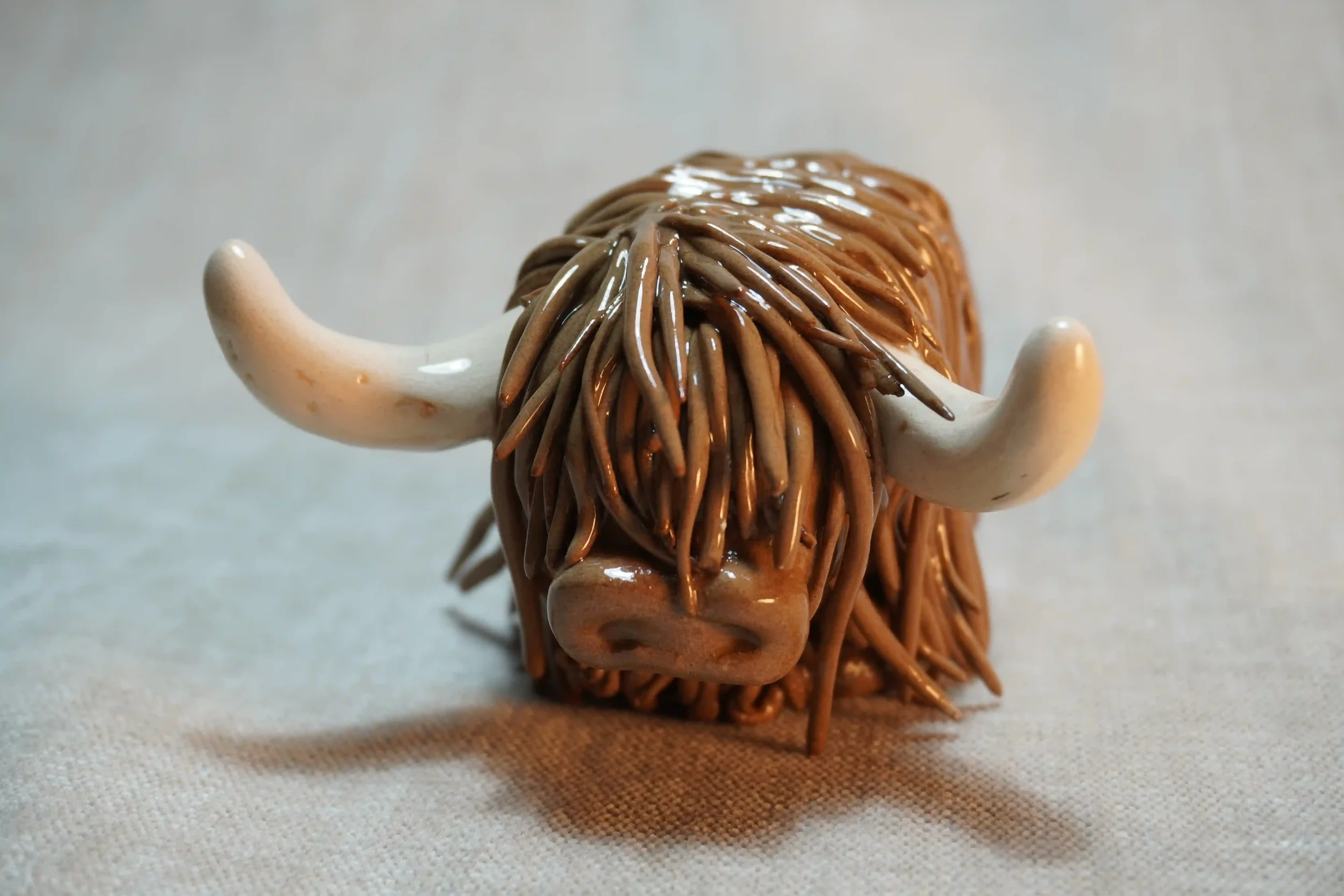 Frontal view of a handmade pottery ceramic sculpture of a Highland Cow ornament with milk chocolate-coloured hair and beige horns.