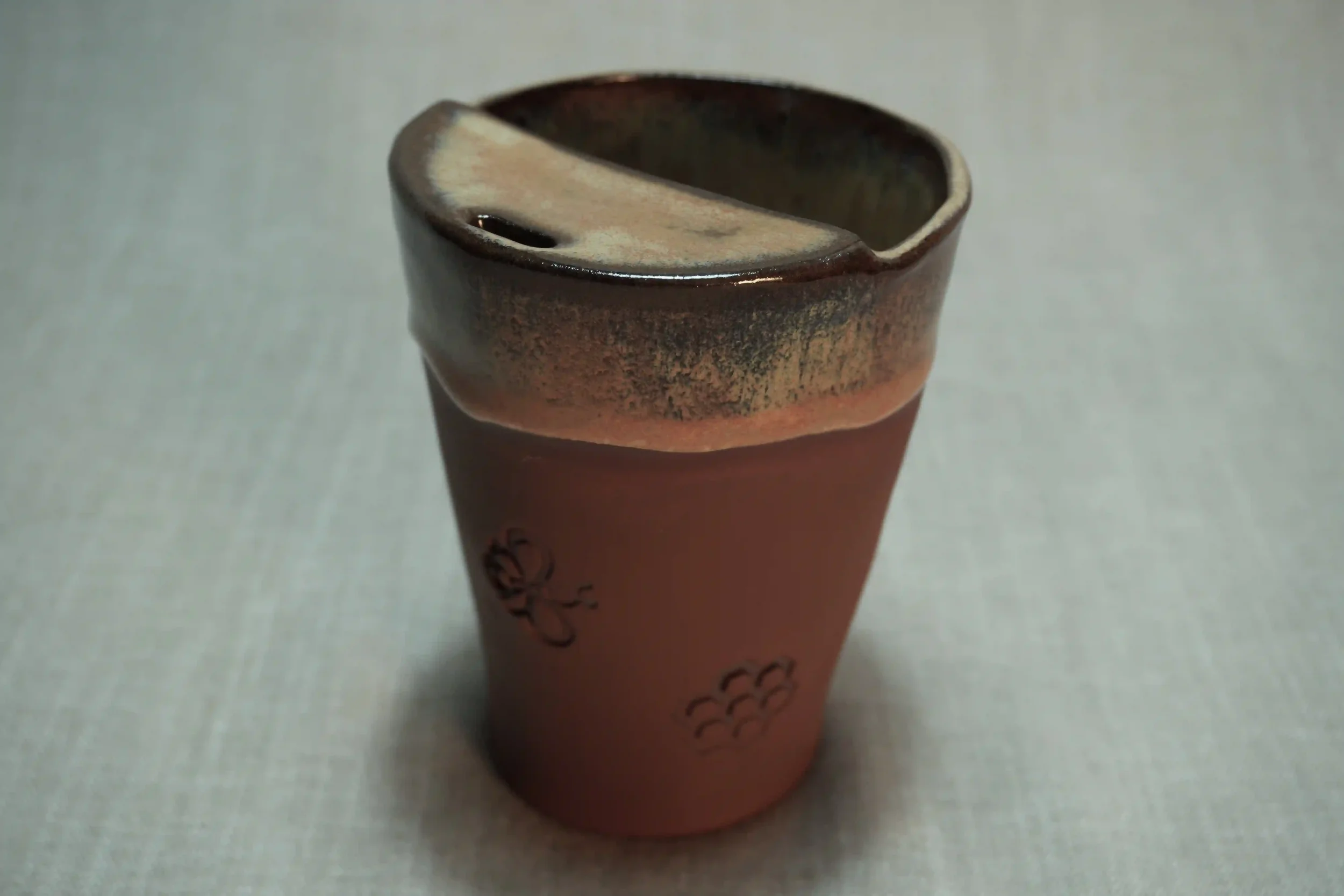 A ceramic travel mug with a honey coloured glaze,  a terracotta brown base, and a decorative bee and honeycomb design on the outside, placed on a light-colored surface.