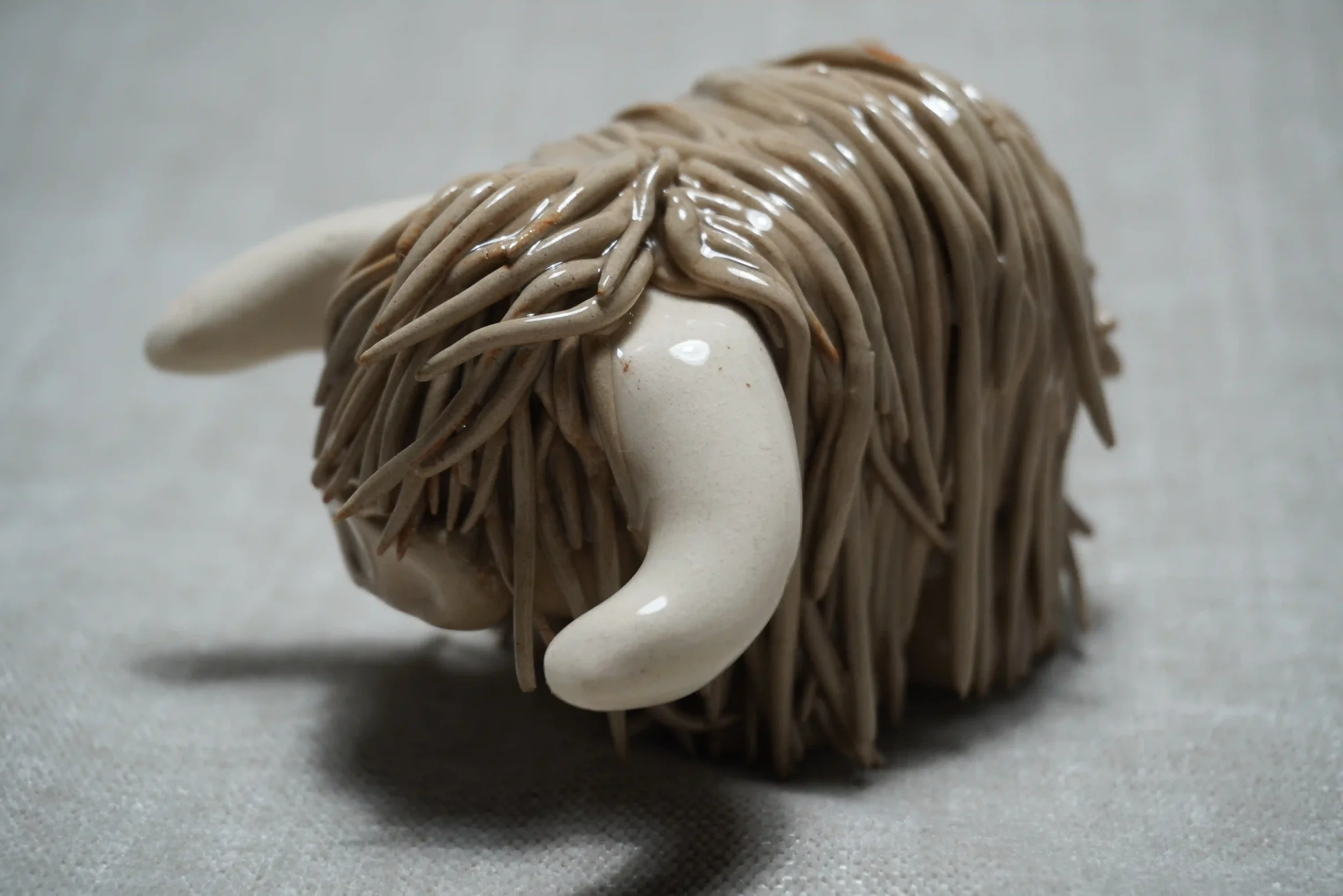 Quarter front view of a cute handmade ceramic Highland Cow ornament with light brown coloured hair and white horns.
