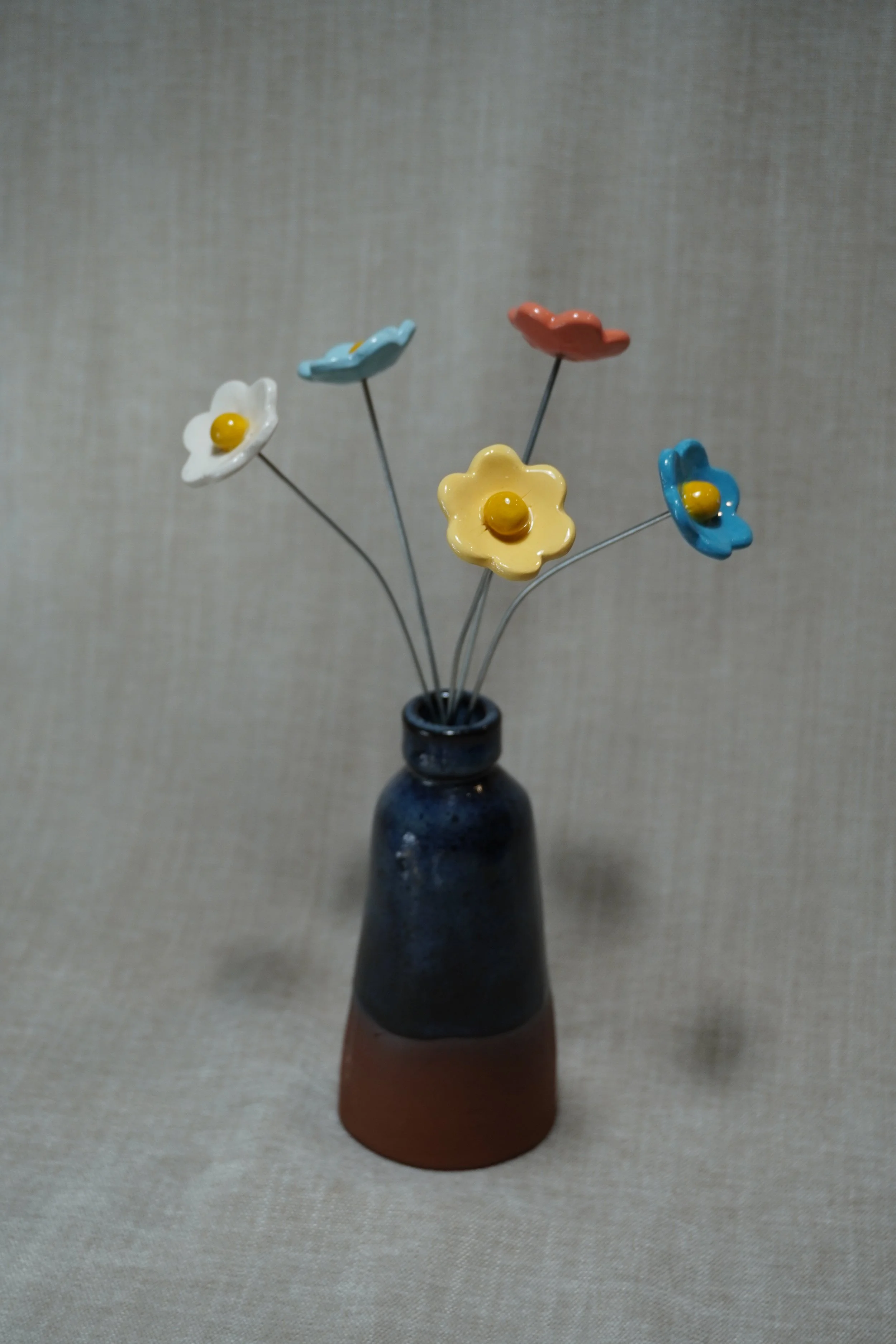 A ceramic pottery vase with a wide, rounded terracotta base, narrow neck, and a blue glaze, holding five ceramic white, yellow, light blue, medium blue, and orange flowers with metal stems, displayed on a light neutral background.