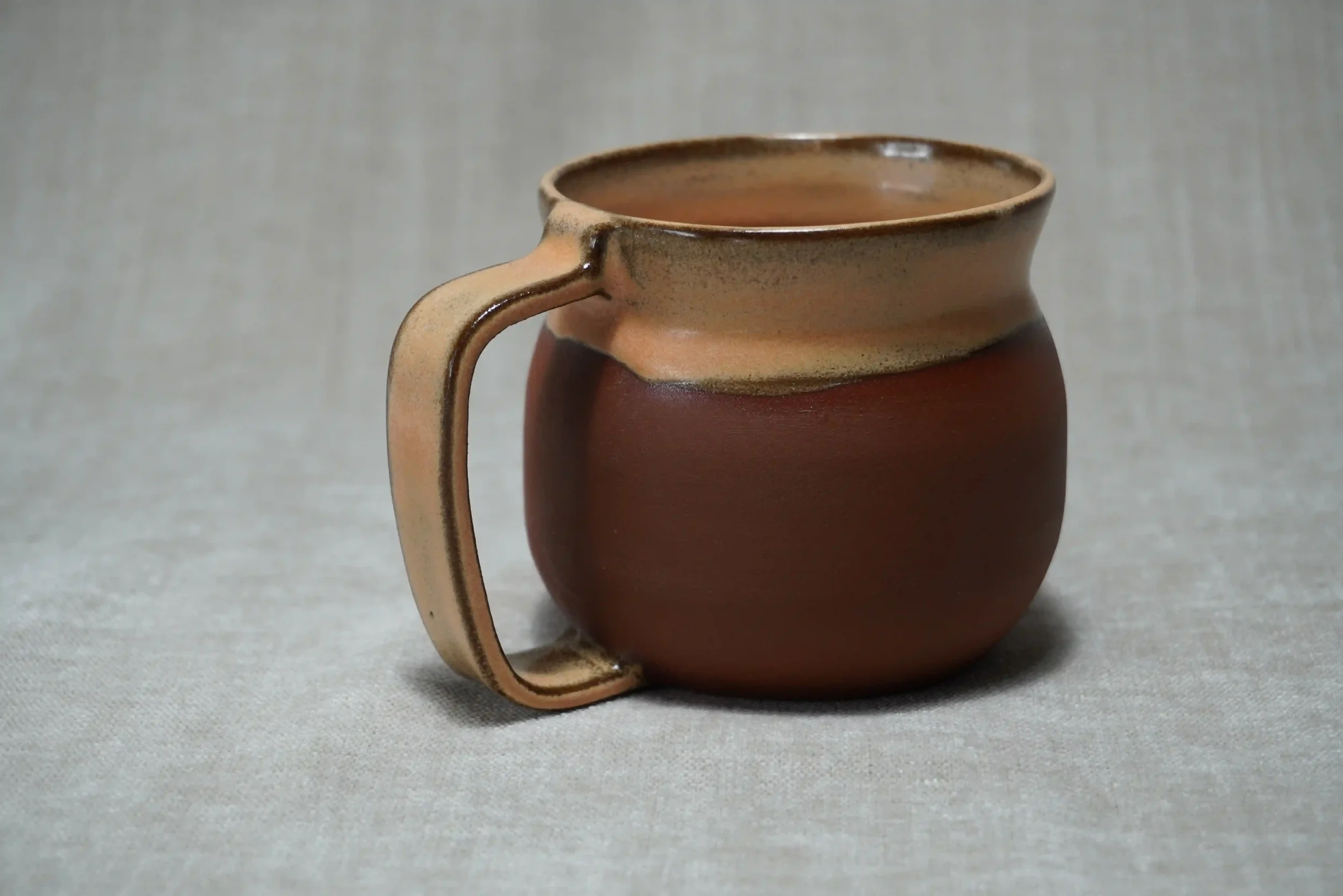 A ceramic pottery mug with a rounded belly shape, a handle on the left, and an orange glaze, placed on a light-colored surface with a neutral background.