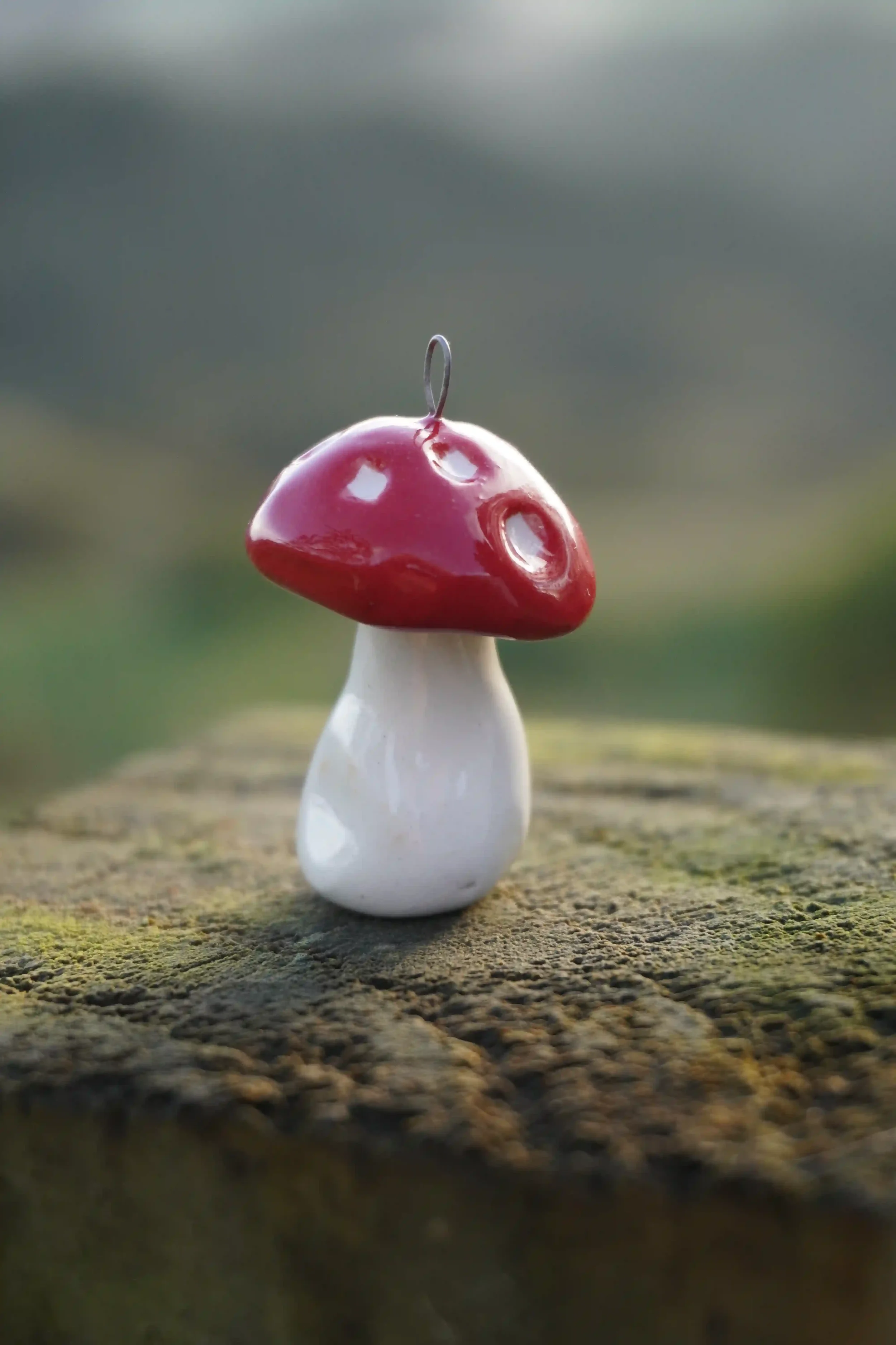 Close-up of a handmade ceramic red and white mushroom figurine with a metal hook on top, placed on a weathered wooden surface outdoors.