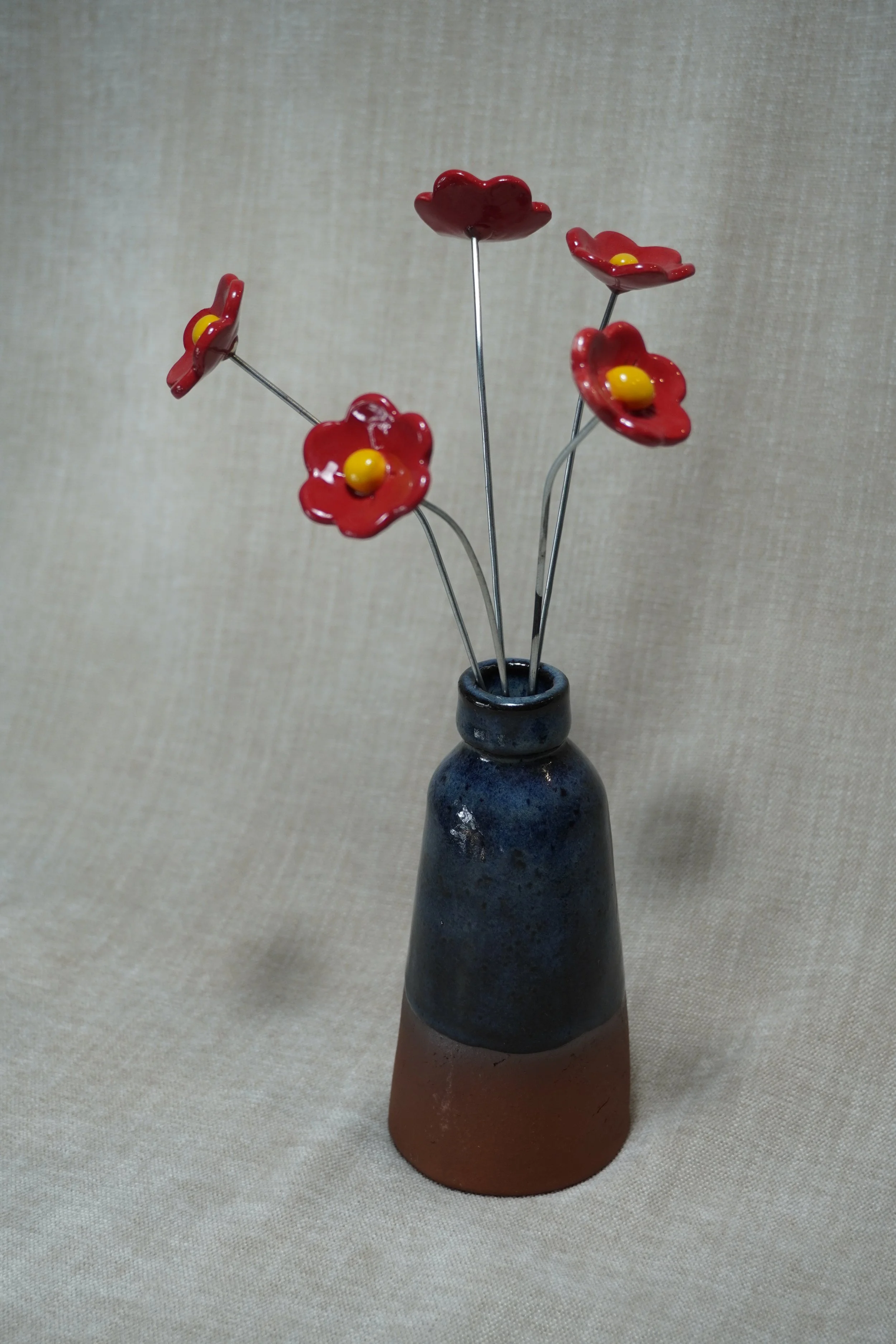 A ceramic vase with a wide, rounded terracotta base, narrow neck, and a blue glaze, holding five red ceramic flowers with yellow centers and metal stems, set against a neutral fabric background.