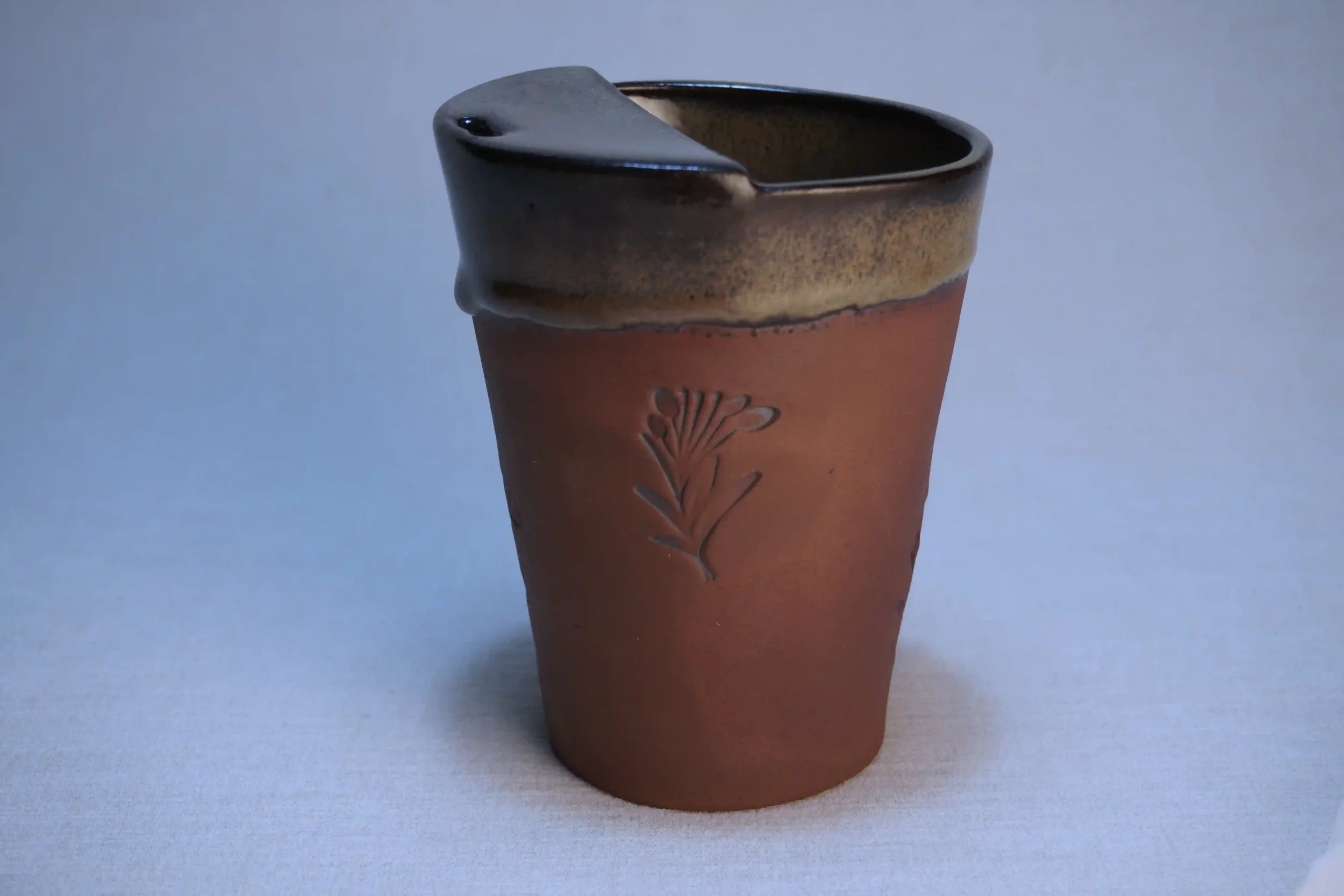 A ceramic cup with a dark glossy interior and a reddish-brown exterior featuring an embossed floral design, set against a plain background.