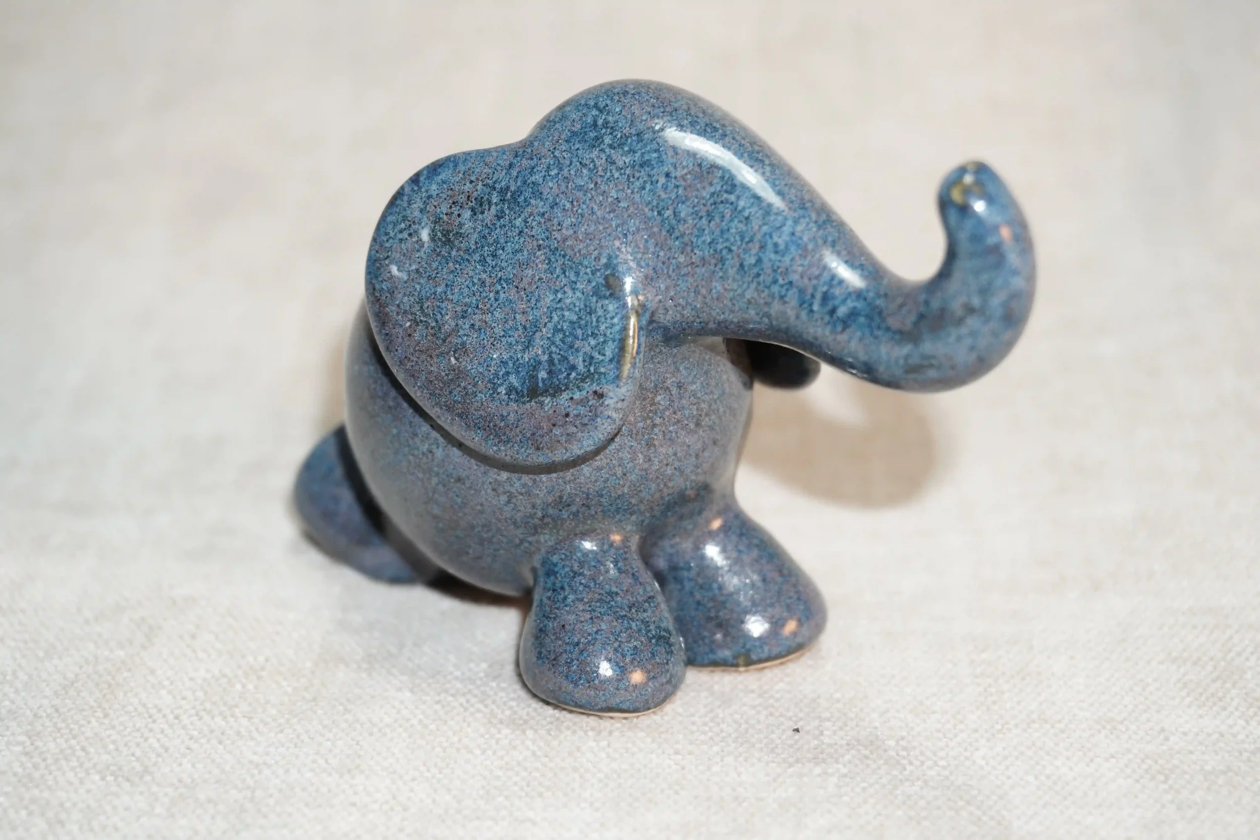 	
	
A small ceramic elephant figurine with a speckled blue glossy glaze finish and it's trunk in the air.